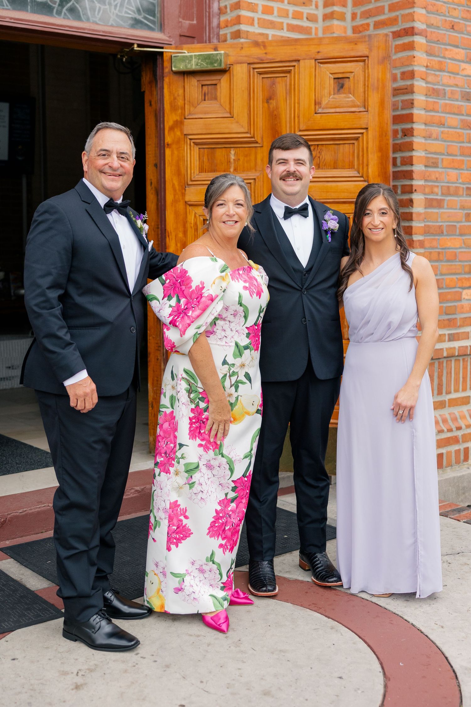 Groom poses with family outside church doors, including parents in formal attire and sister in lavender bridesmaid dress.