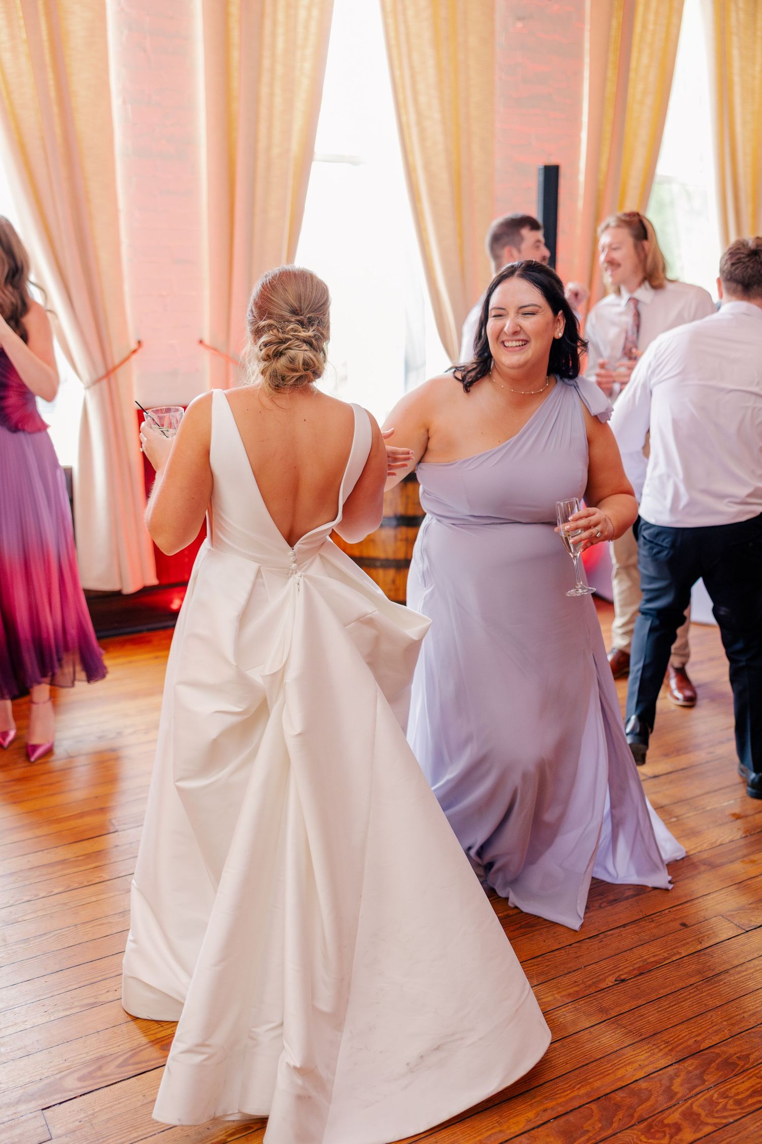Bride in white gown dancing with bridesmaid in lavender dress at elegant wedding reception.