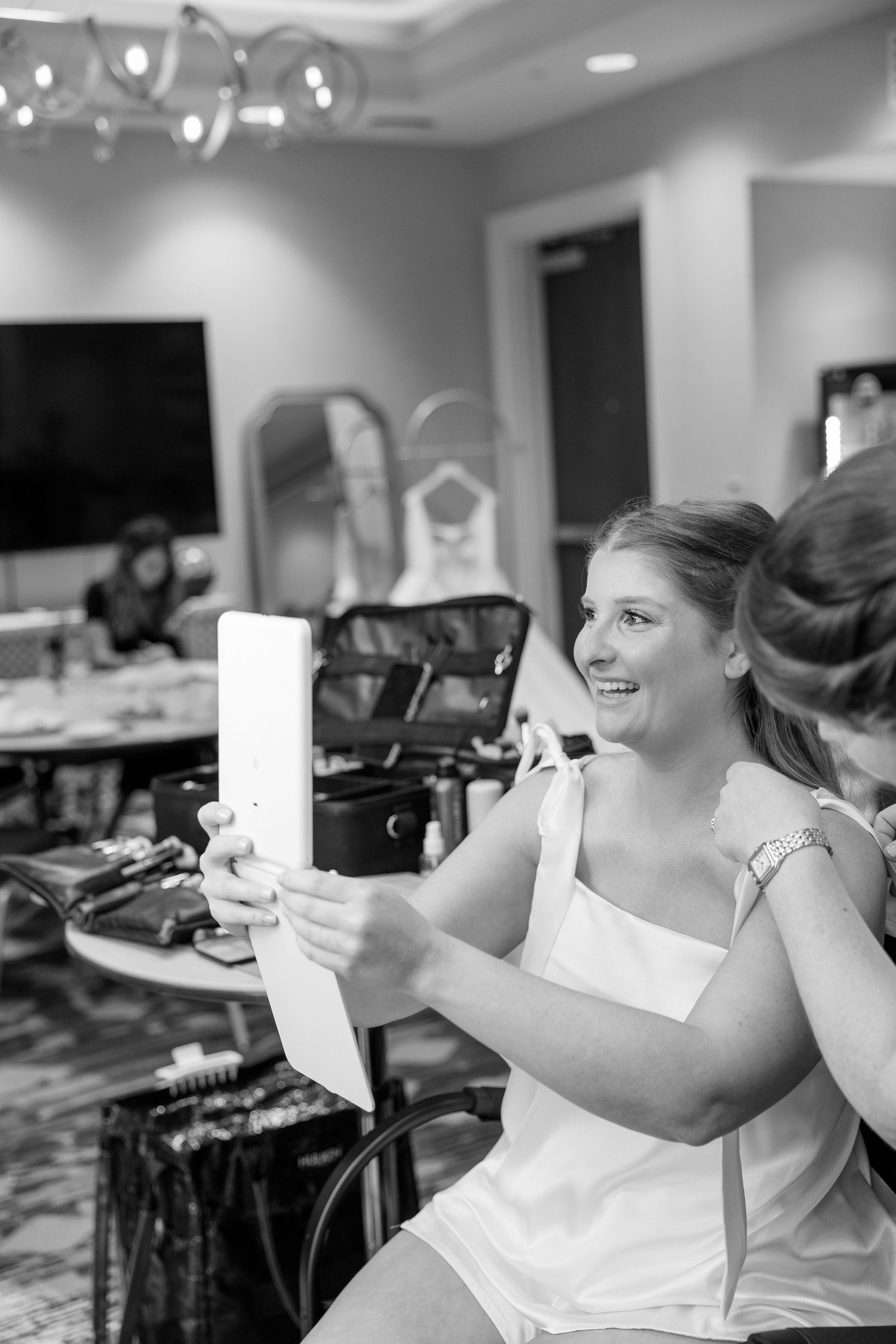 Black and white photo of bride smiling while getting makeup done, holding mirror during wedding day preparations.