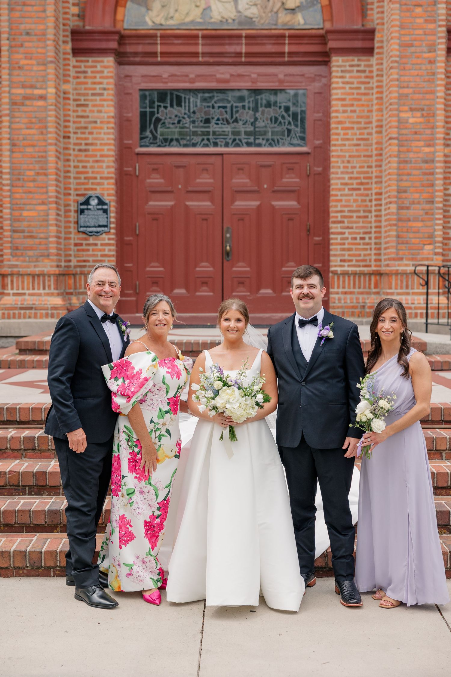 Bride and groom with family members posing on church steps in front of red wooden doors.