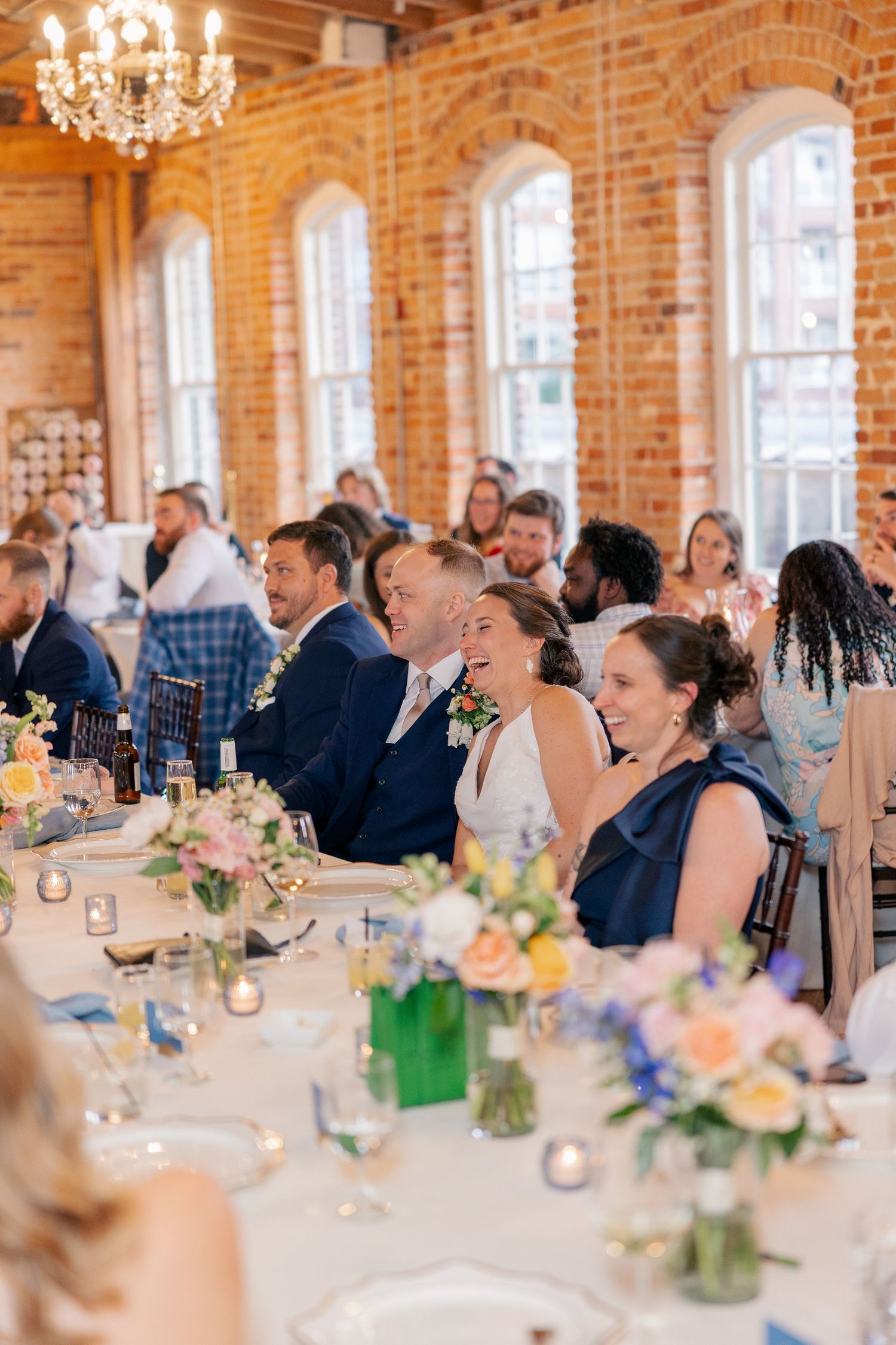 Bride and groom smile during wedding reception speeches in elegant brick-walled banquet hall.