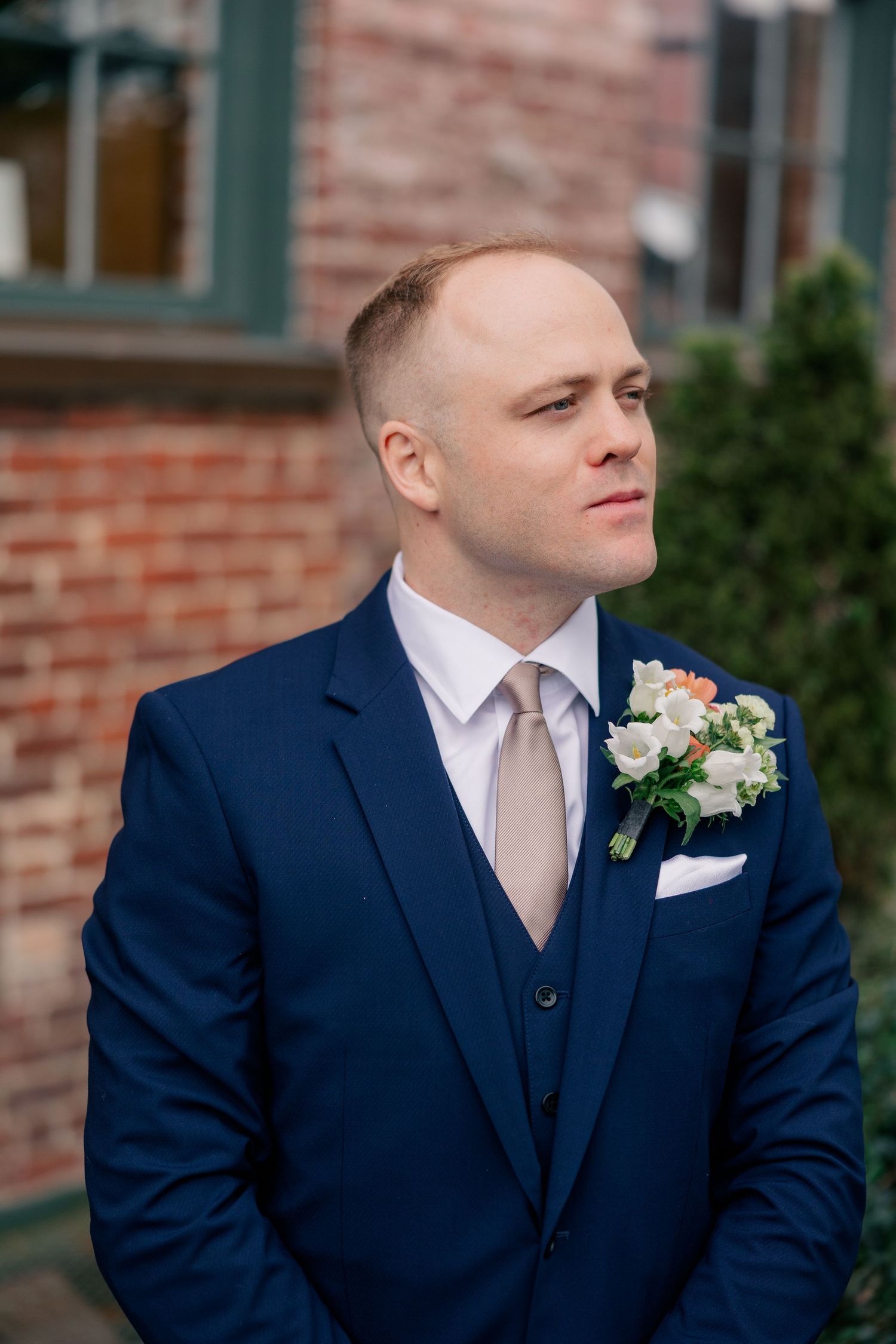 Handsome groom in navy suit with floral boutonniere gazes thoughtfully outside brick building.