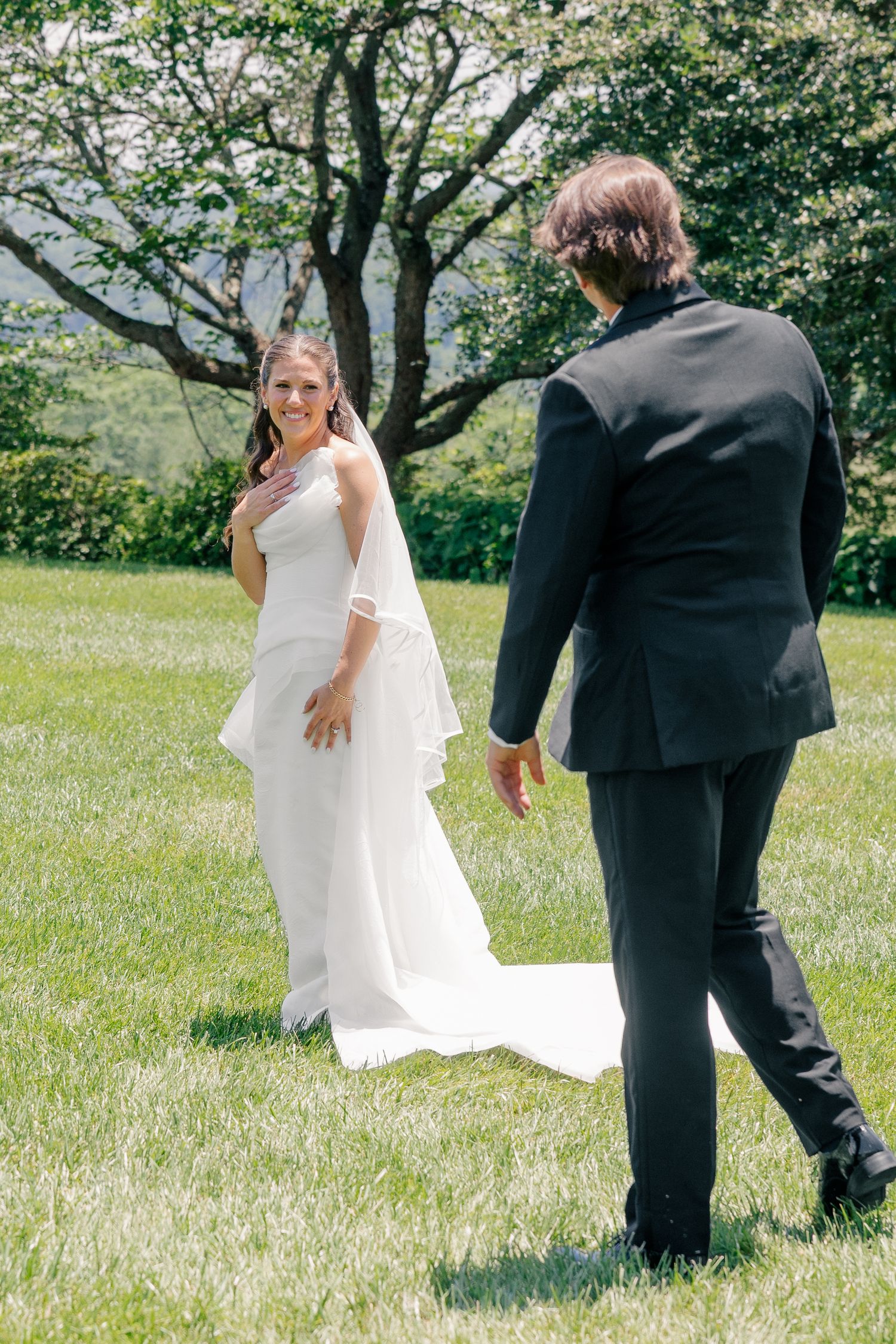 Bride smiling at groom approaching her during outdoor first look on sunny green lawn.