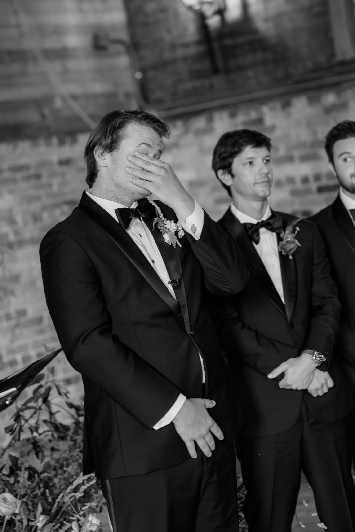 Emotional groom covering his face at the altar in black tuxedo as groomsmen stand behind him.