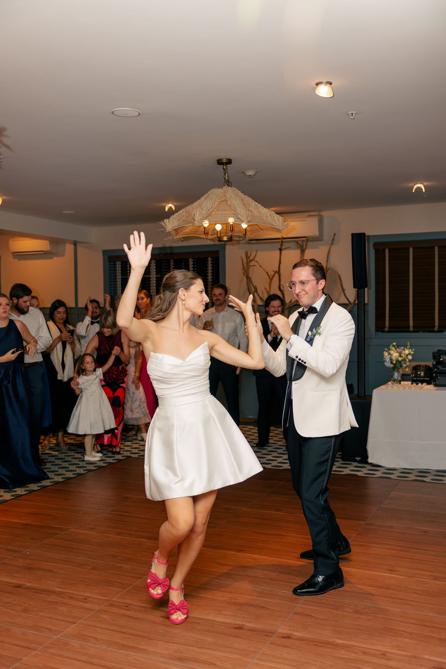 Bride in short white dress and pink heels dances with groom in white tuxedo at wedding reception.