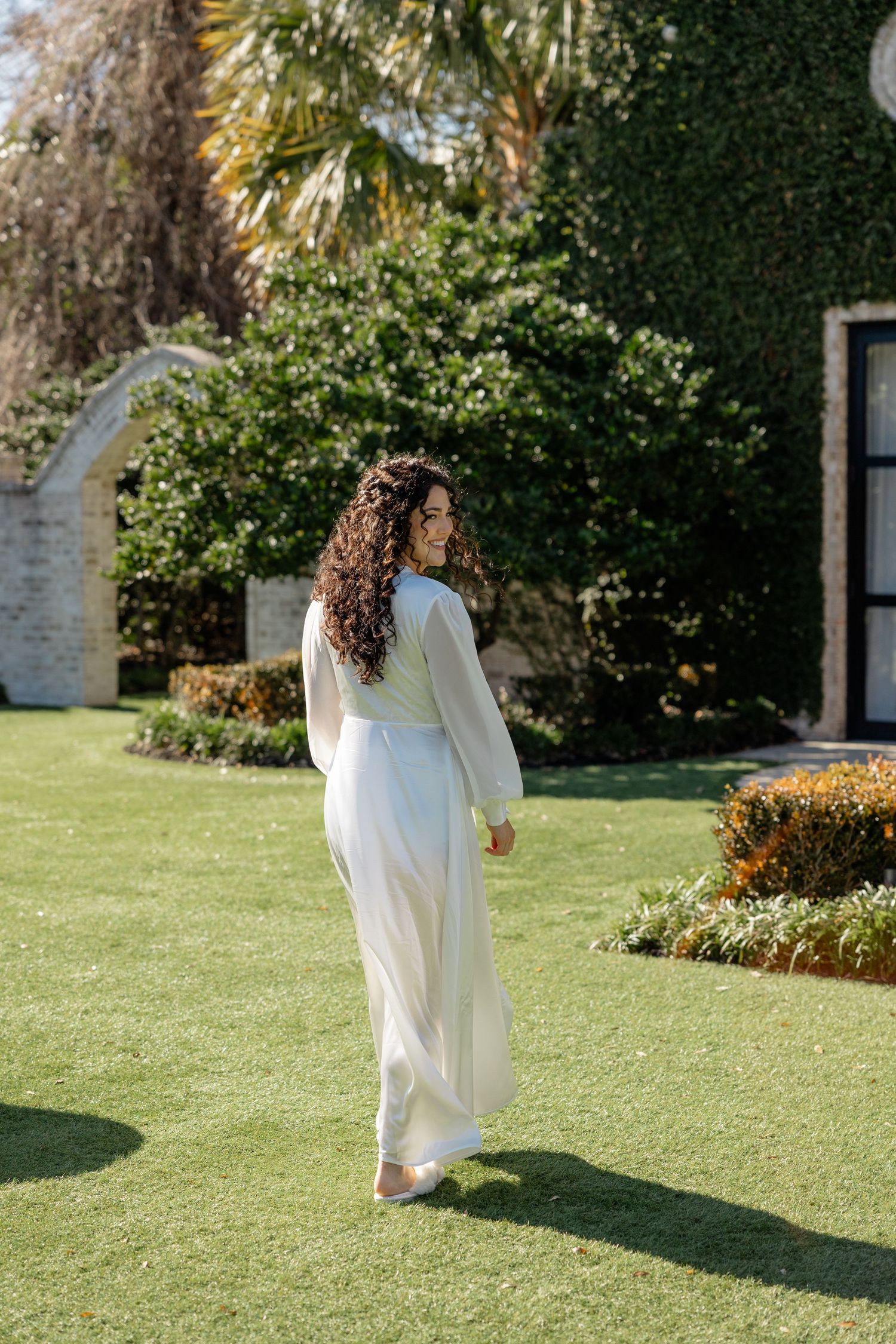 Woman in flowing white dress walking through a lush garden setting on a sunny day.