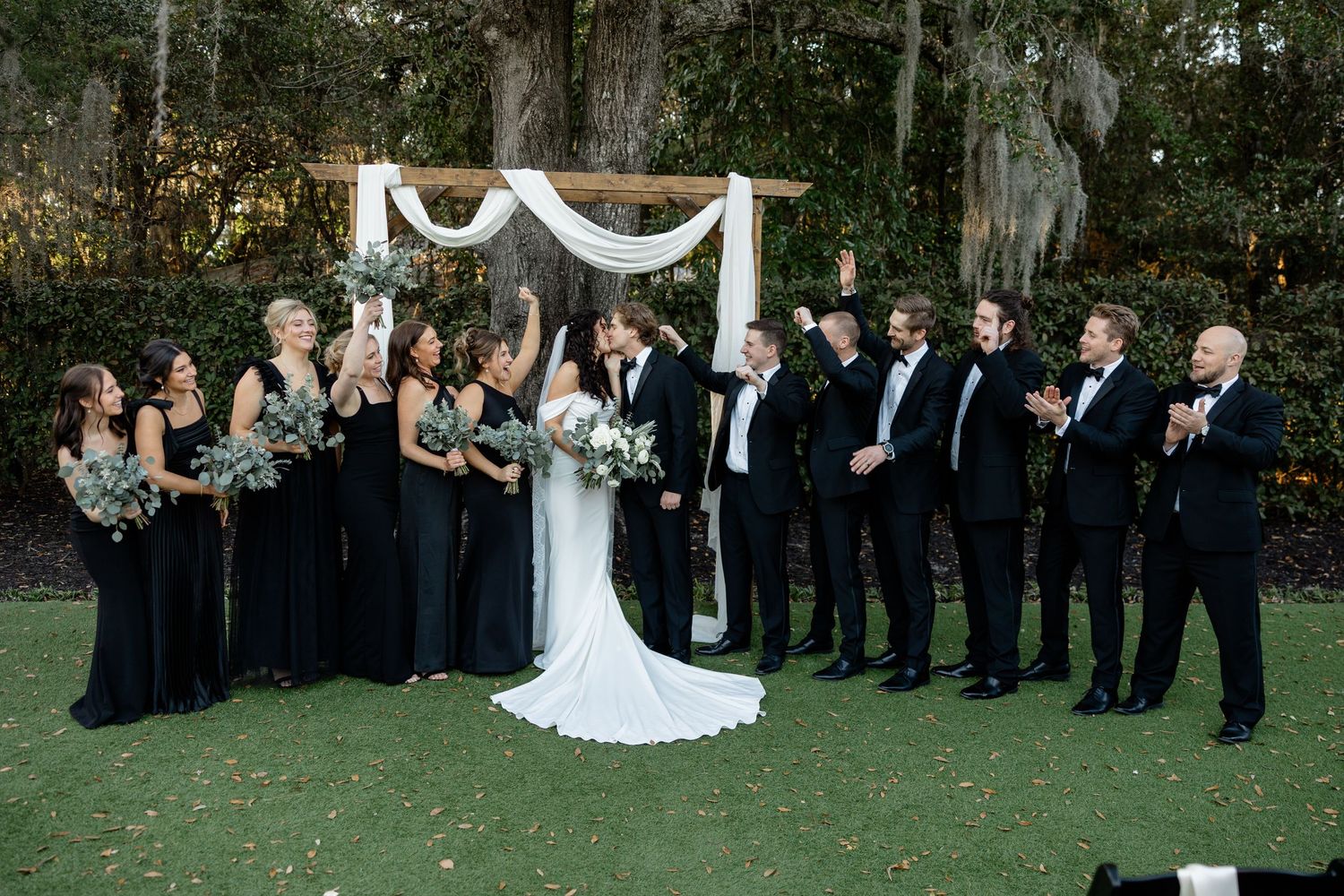A wedding party in black and white attire poses together under a decorated arch.