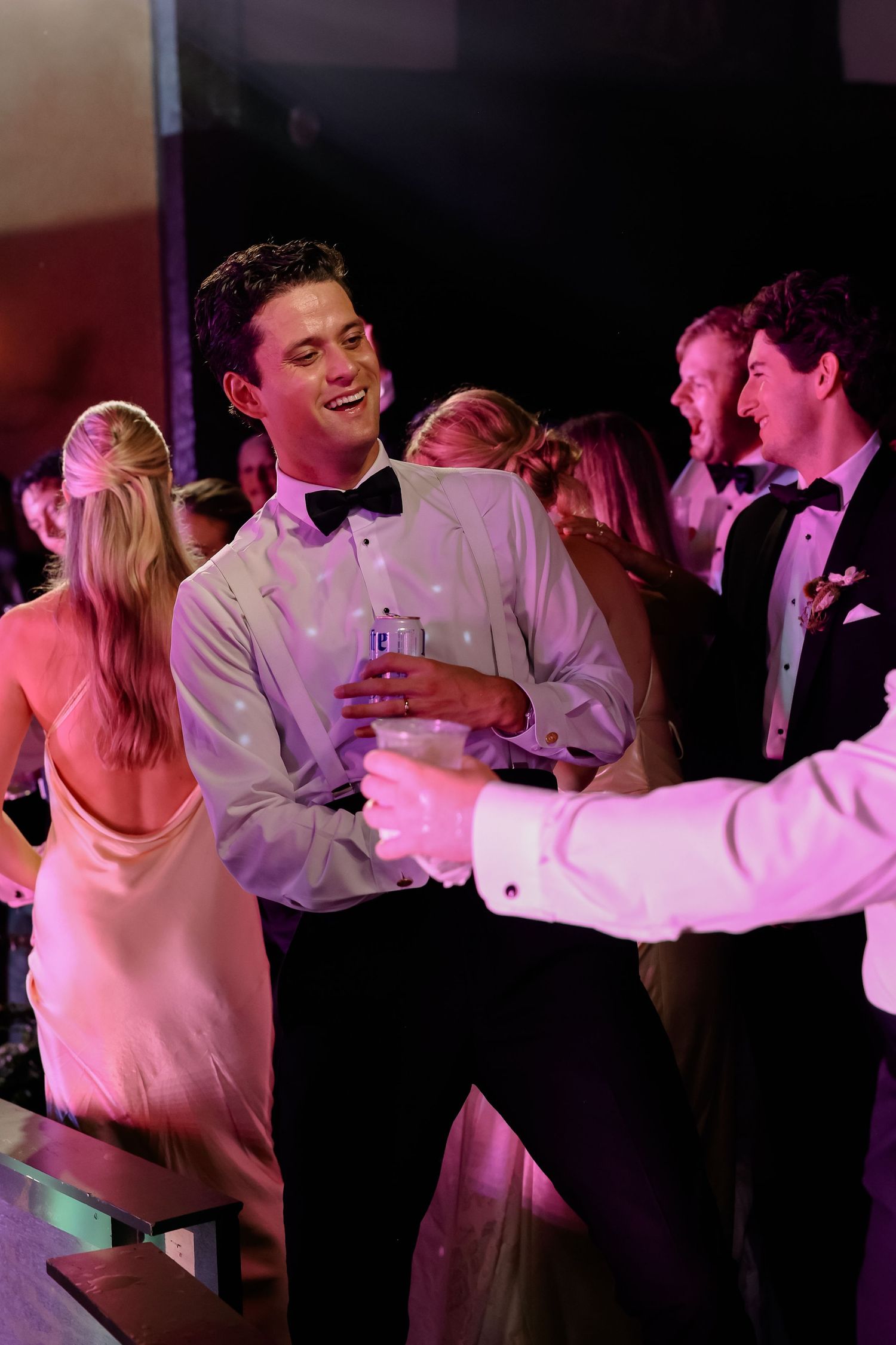 Guests in formal attire dance and celebrate joyfully at a wedding reception under pink lighting.
