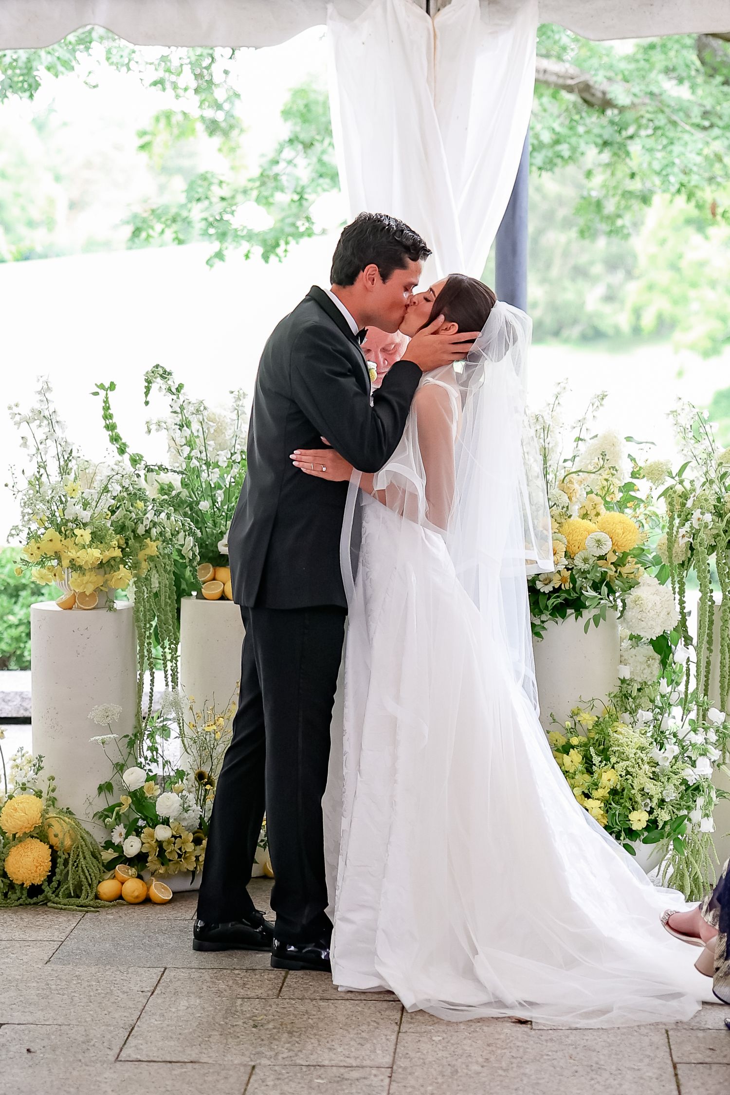 A bride and groom share their first kiss at a wedding ceremony surrounded by yellow and white floral arrangements.