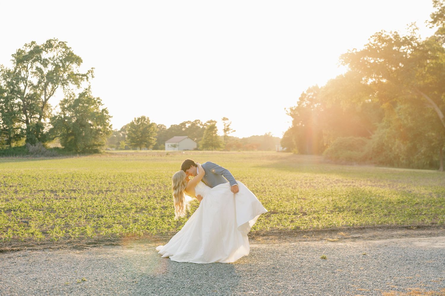 Bride and groom share a dip kiss bathed in warm sunset light on a rustic farm path.