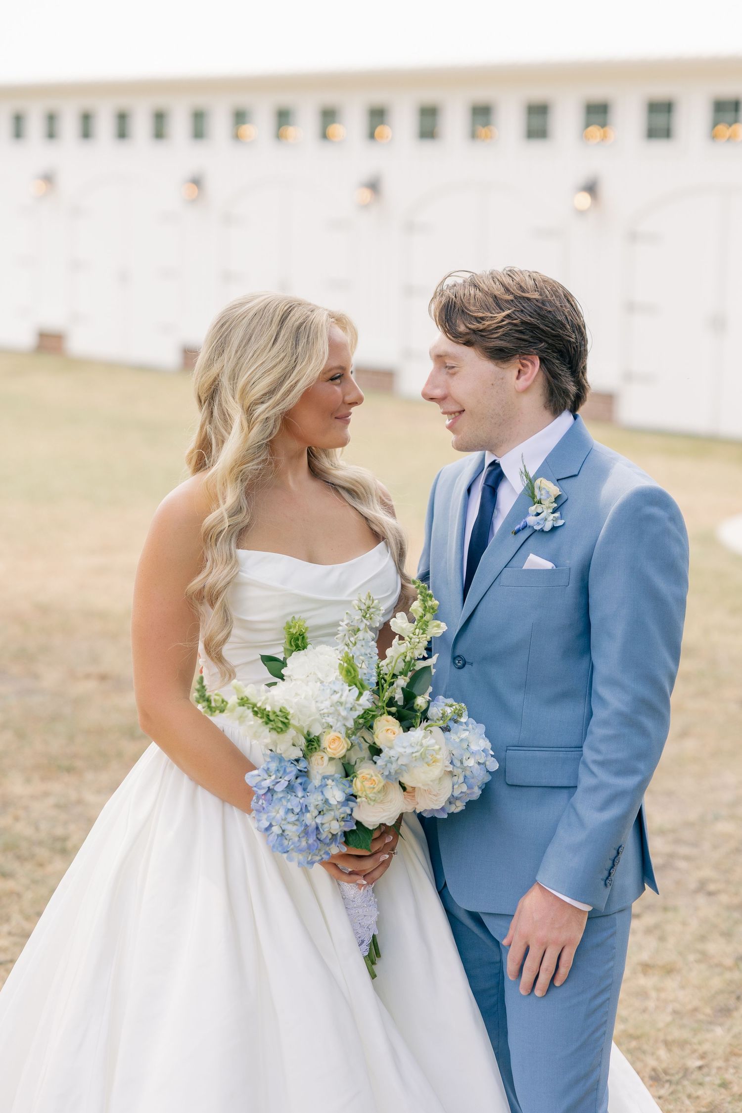 Bride and groom gazing at each other, bride holding blue and white floral bouquet at farm wedding venue.