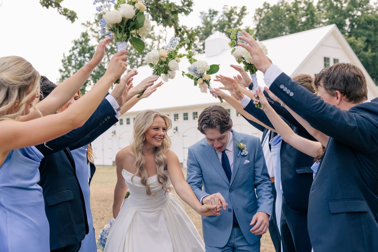 Bride and groom walk through an arch of raised bouquets held by wedding party in blue attire outside a white barn.
