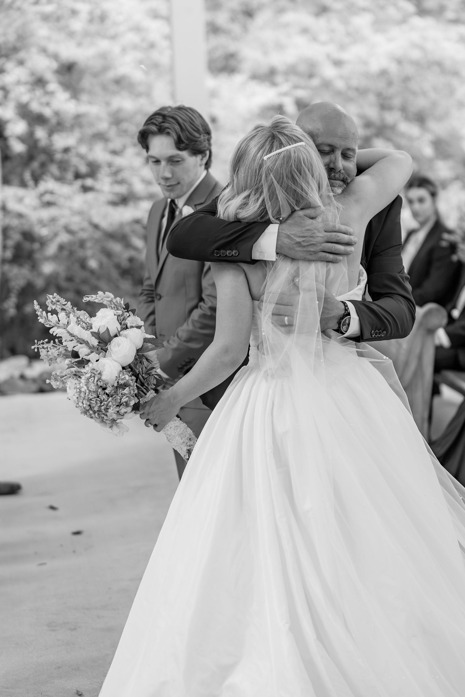 Black and white photo of bride hugging man at altar while groom stands beside her during wedding ceremony.