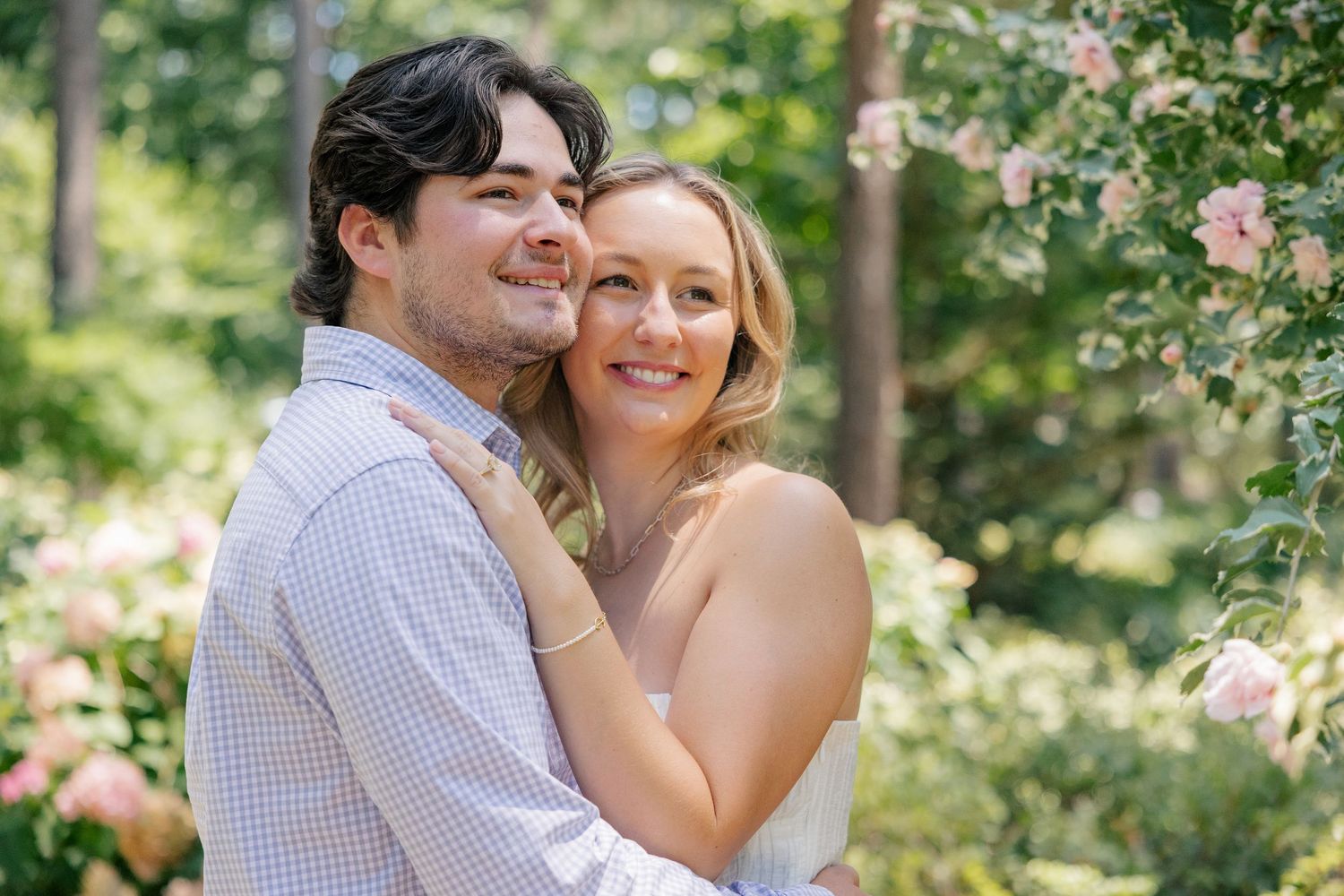 Happy engaged couple embracing among blooming pink roses in lush garden setting.
