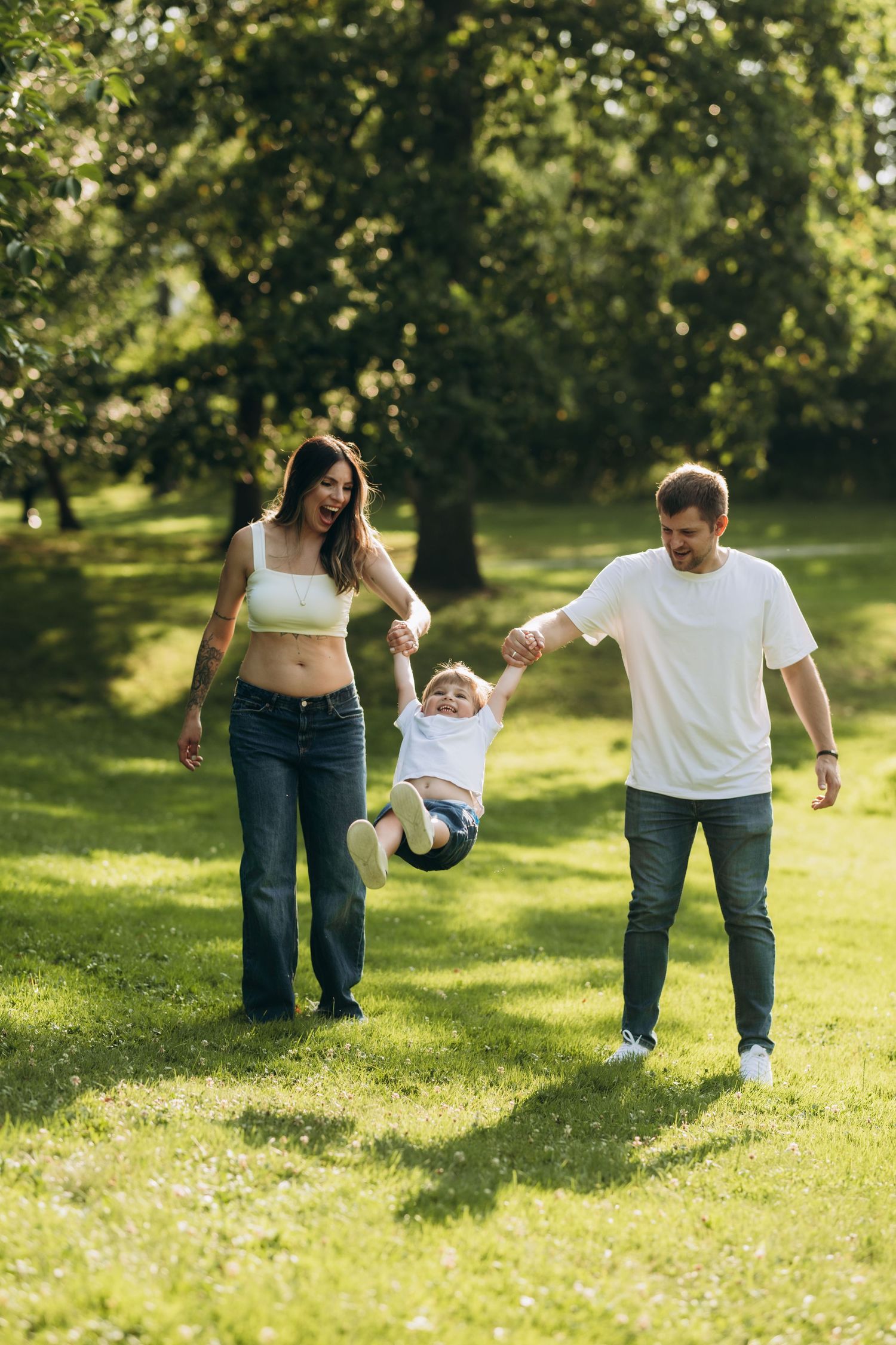 A family enjoys a playful moment swinging their small child between them on a sunny day in a lush green park.