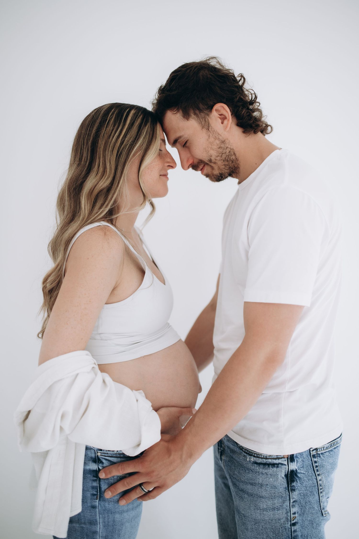 A couple in white tops and jeans embraces intimately against a light backdrop in a romantic maternity photoshoot.