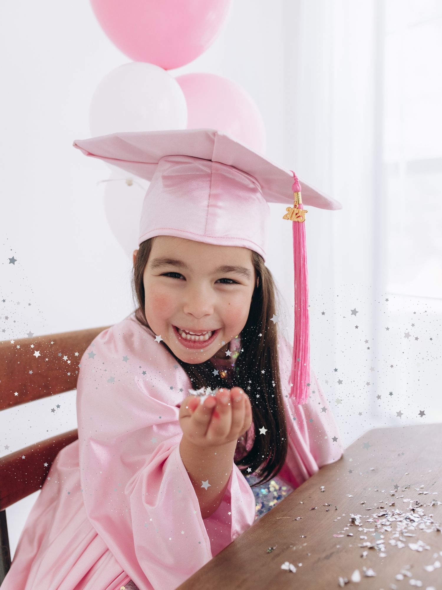 A graduate in pink cap and gown throws confetti while celebrating with a pink balloon in a bright room.