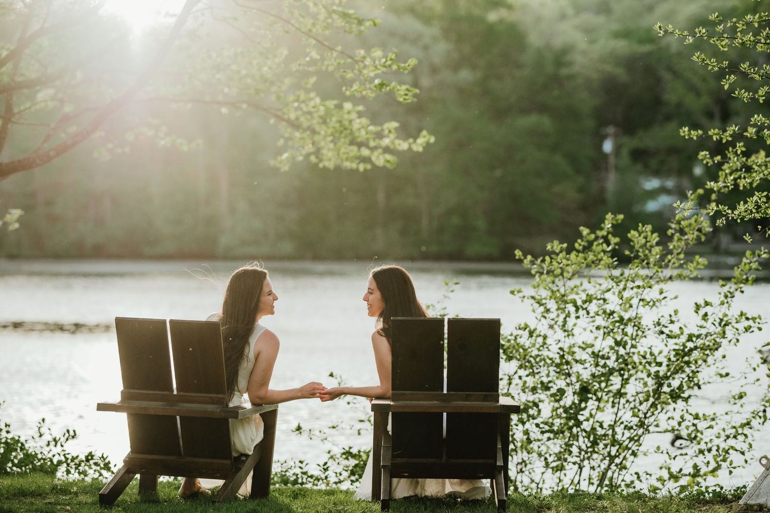Two people relaxing in Adirondack chairs by a lake during golden hour.