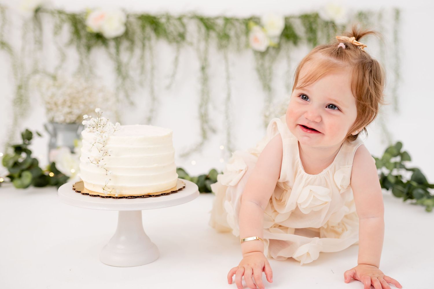 Baby in cream dress crawls near white cake with floral garland backdrop.