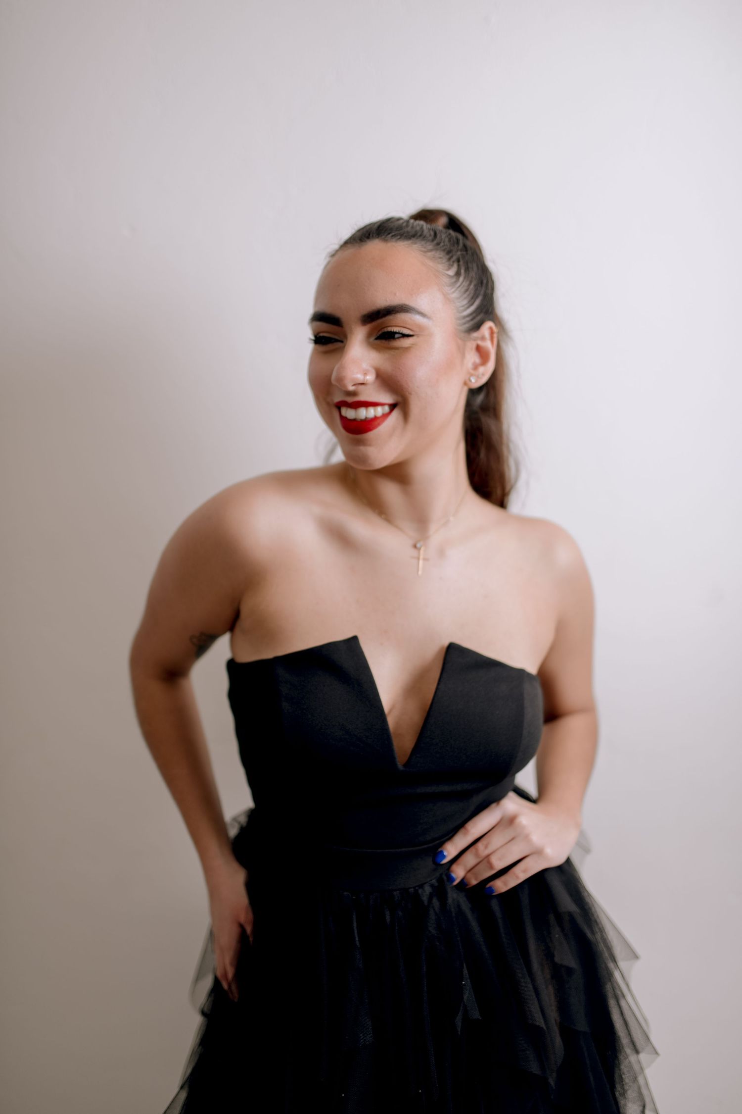 A woman in a black strapless dress poses with a bright smile against a white wall.