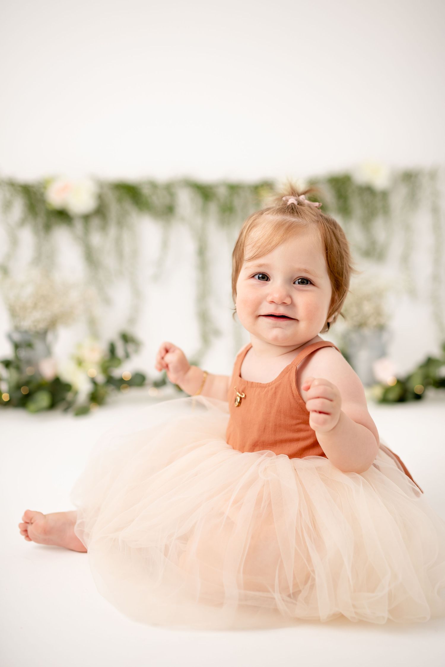 Baby in peach tulle dress sits against floral backdrop during portrait session.