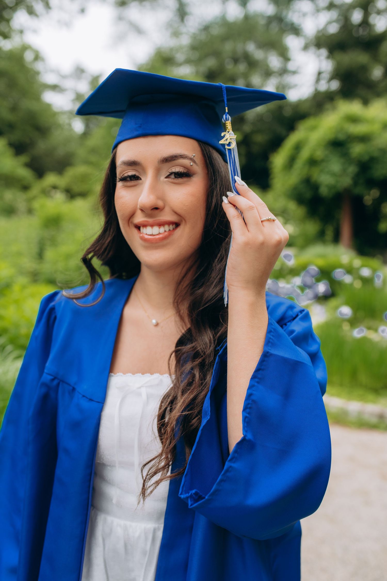 Graduate wearing blue cap and gown smiles while touching tassel during outdoor graduation photoshoot.