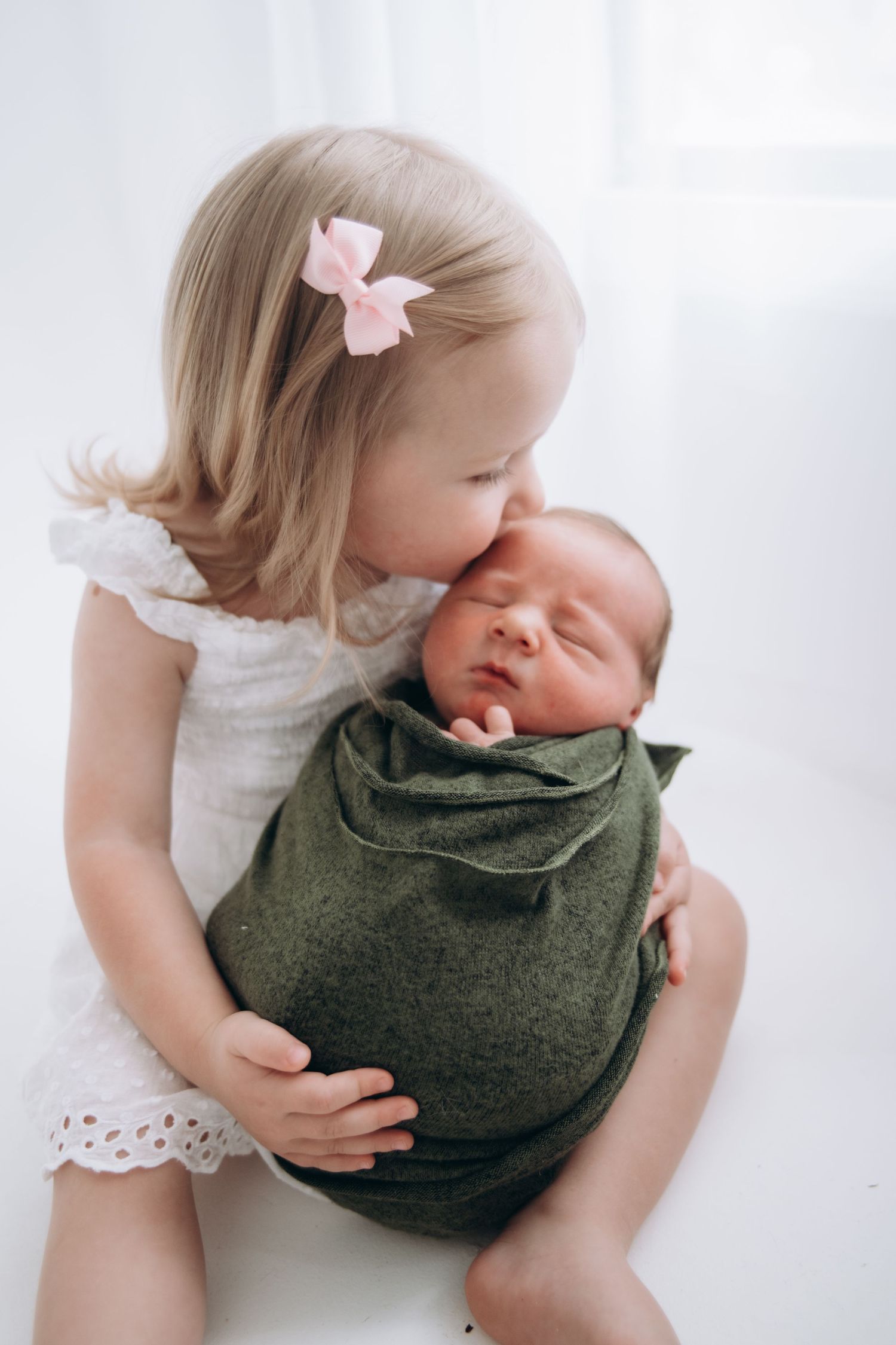 A tender sibling moment shows a young child in a white outfit embracing a swaddled baby in an olive green blanket.