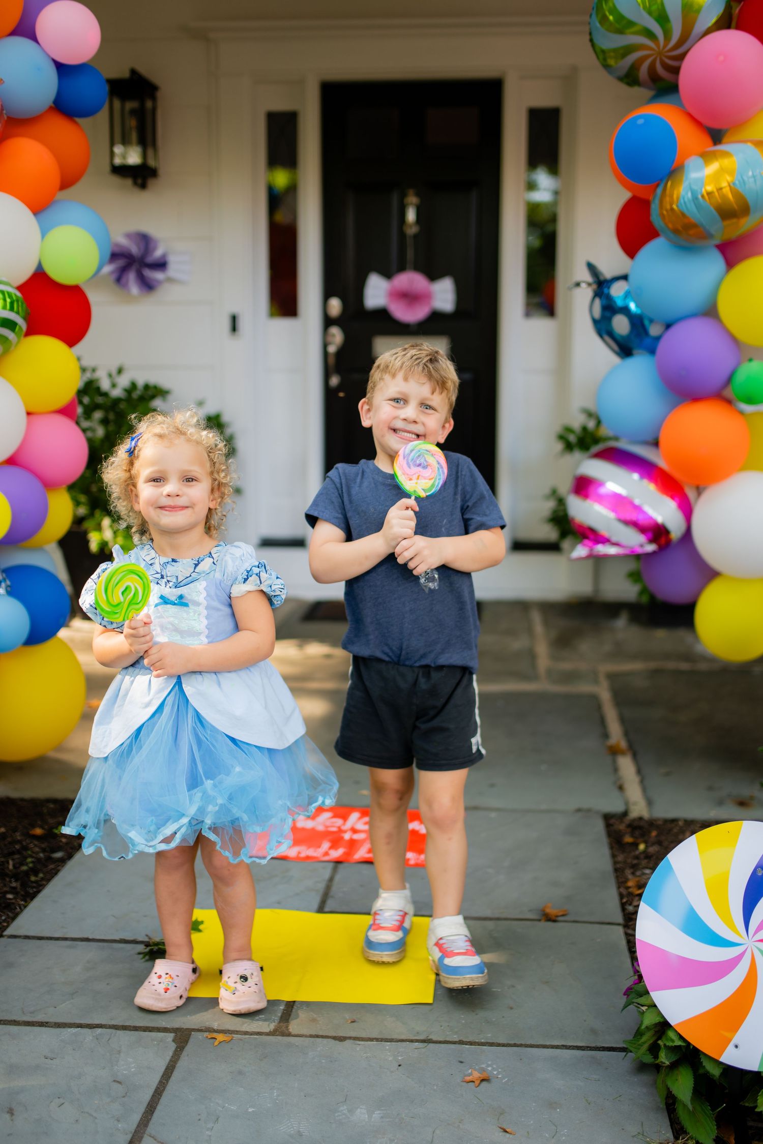 Children holding lollipops in front of house decorated with colorful balloon arch.