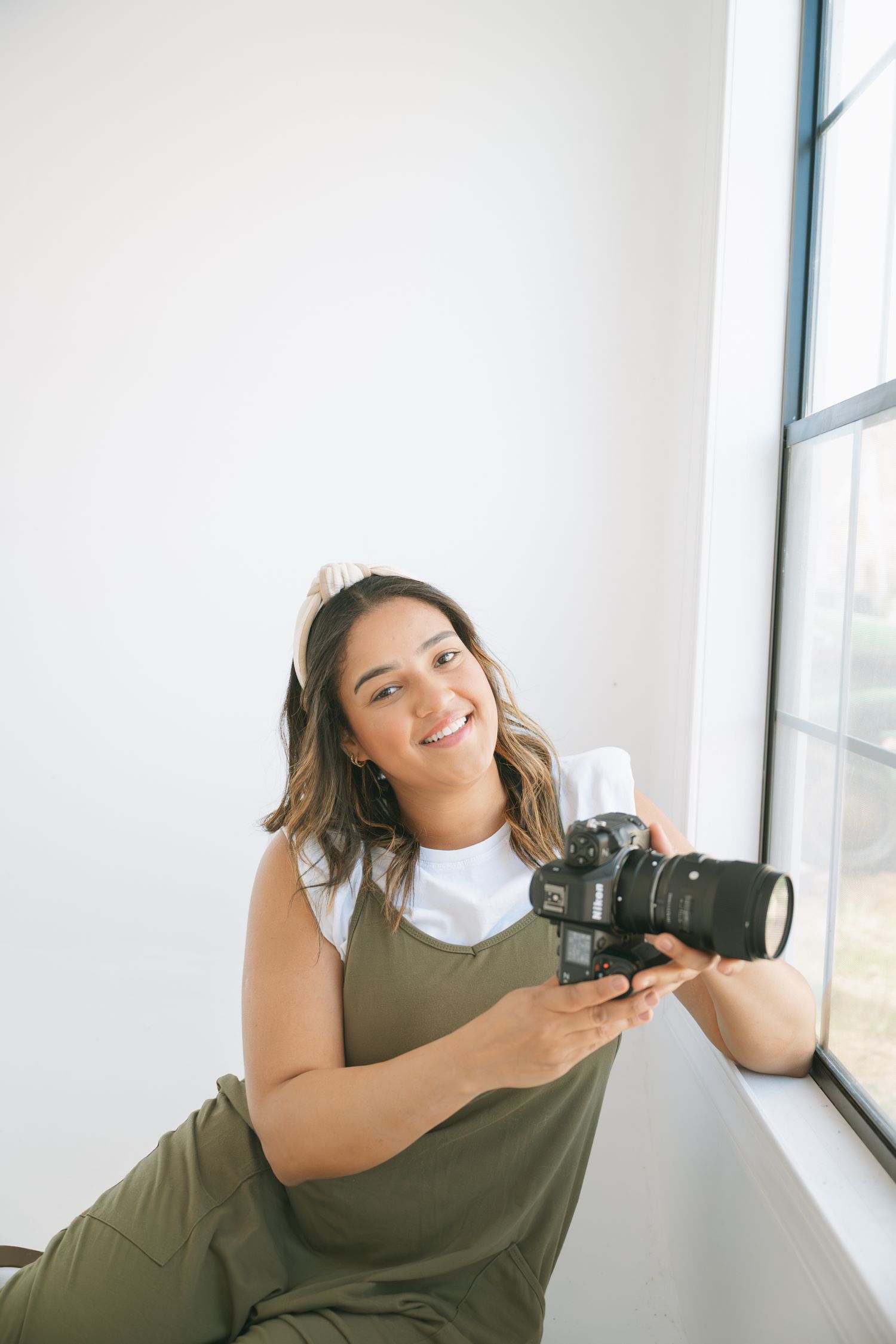 Photographer in olive green overalls sits by window holding professional camera with telephoto lens.