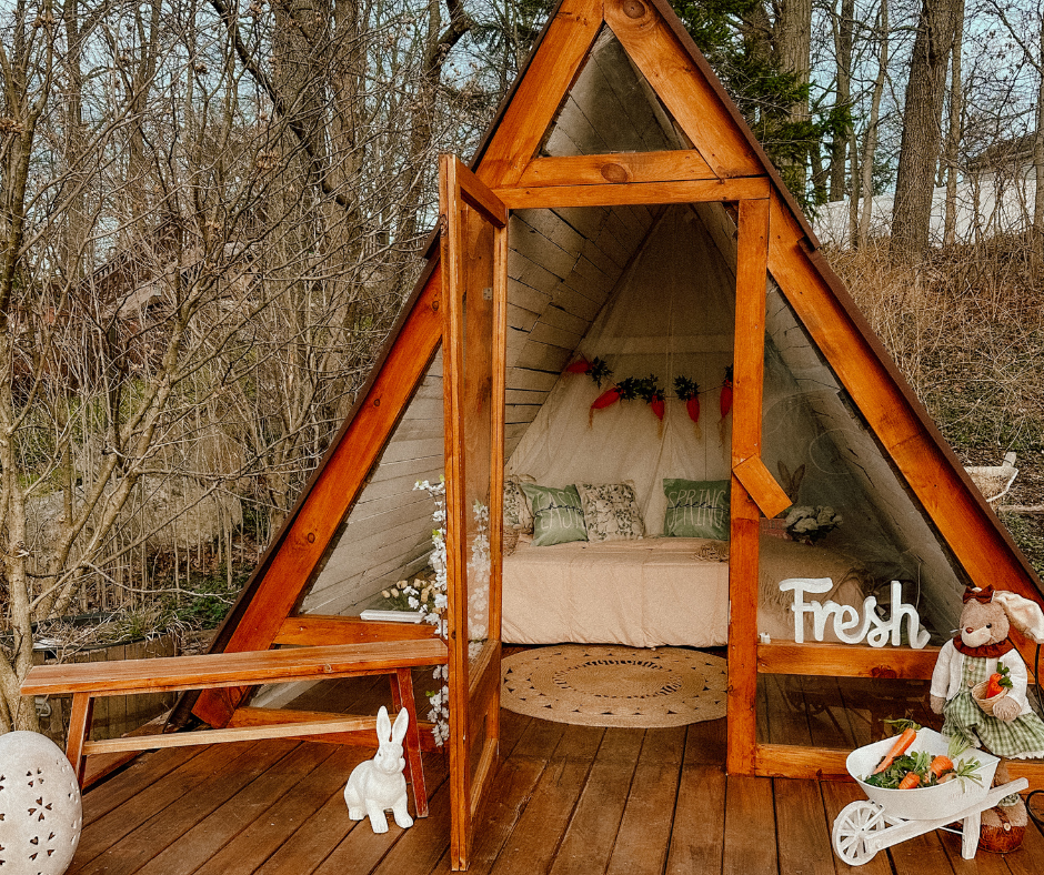 A cozy A-frame cabin with a wooden deck features warm lighting and plush bedding in a forest setting.
