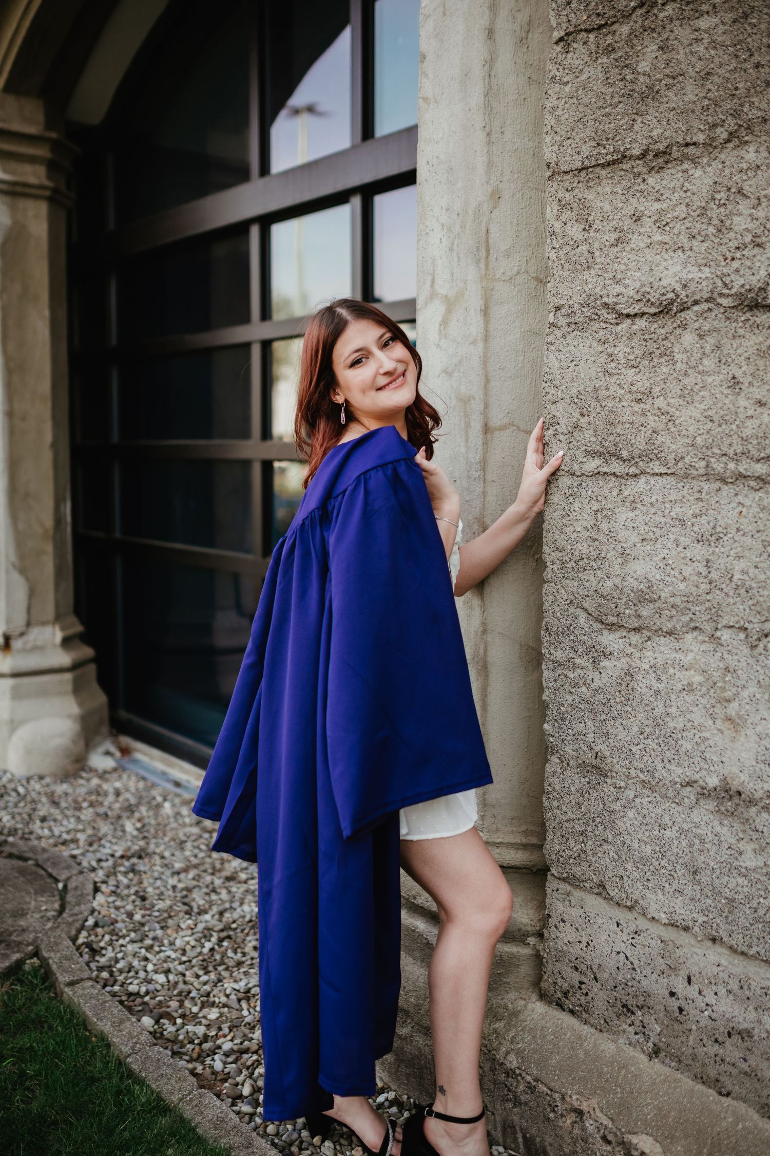 Graduate in blue graduation gown poses against stone column outdoors.