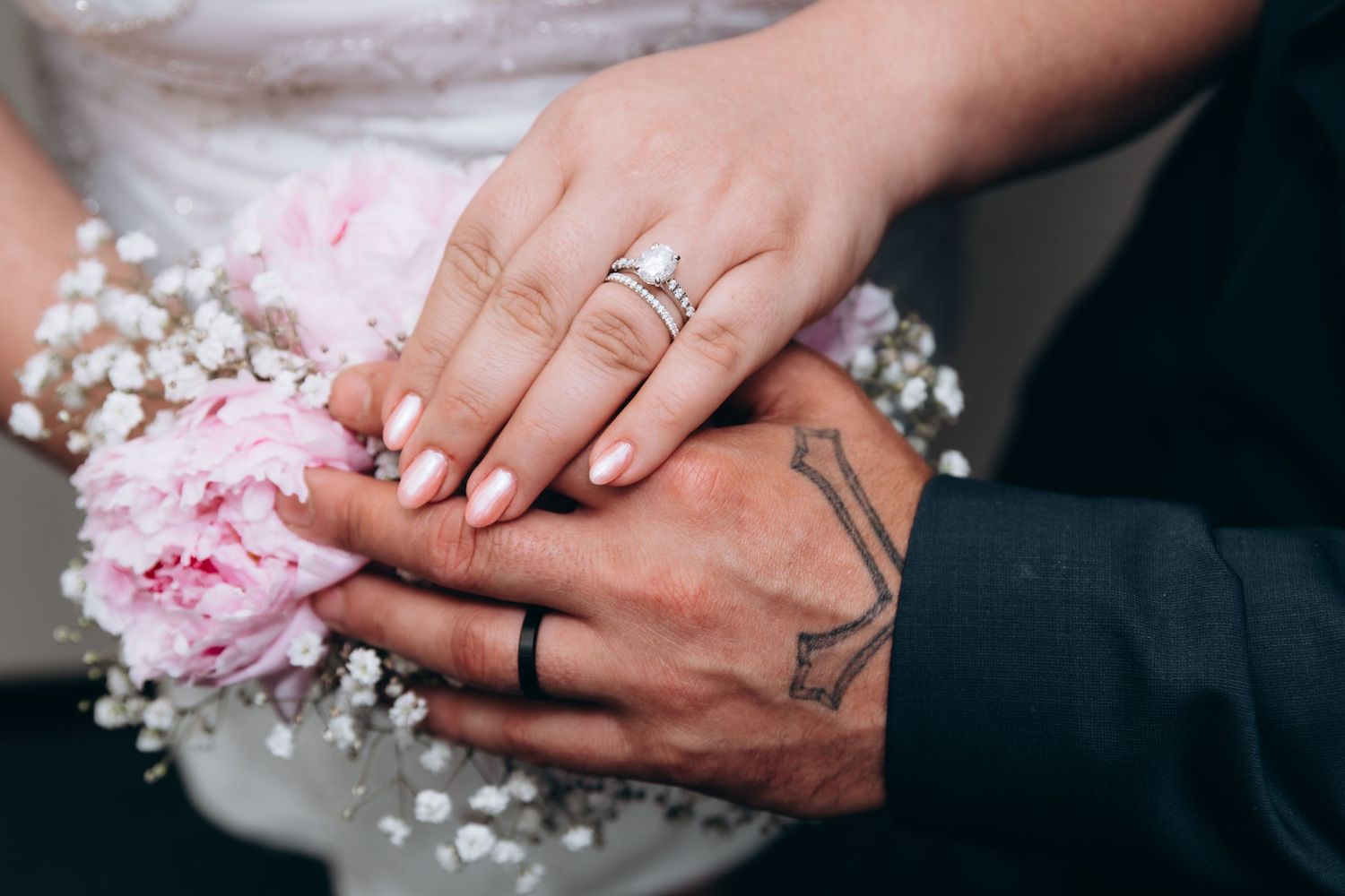 A close-up of wedding hands with rings, delicate pink peony flower and baby's breath florals in the background.