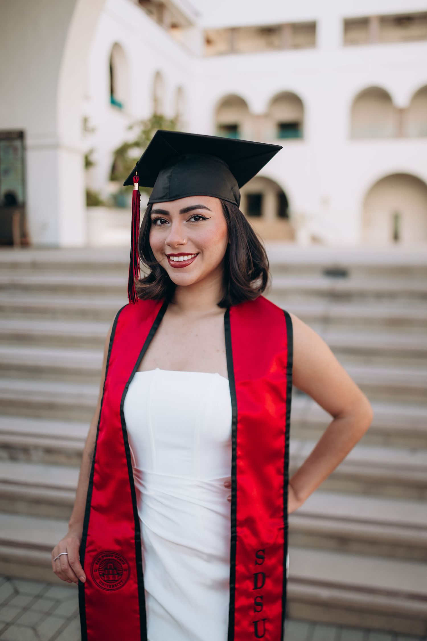 Graduate in white dress and red stole smiling on campus steps.