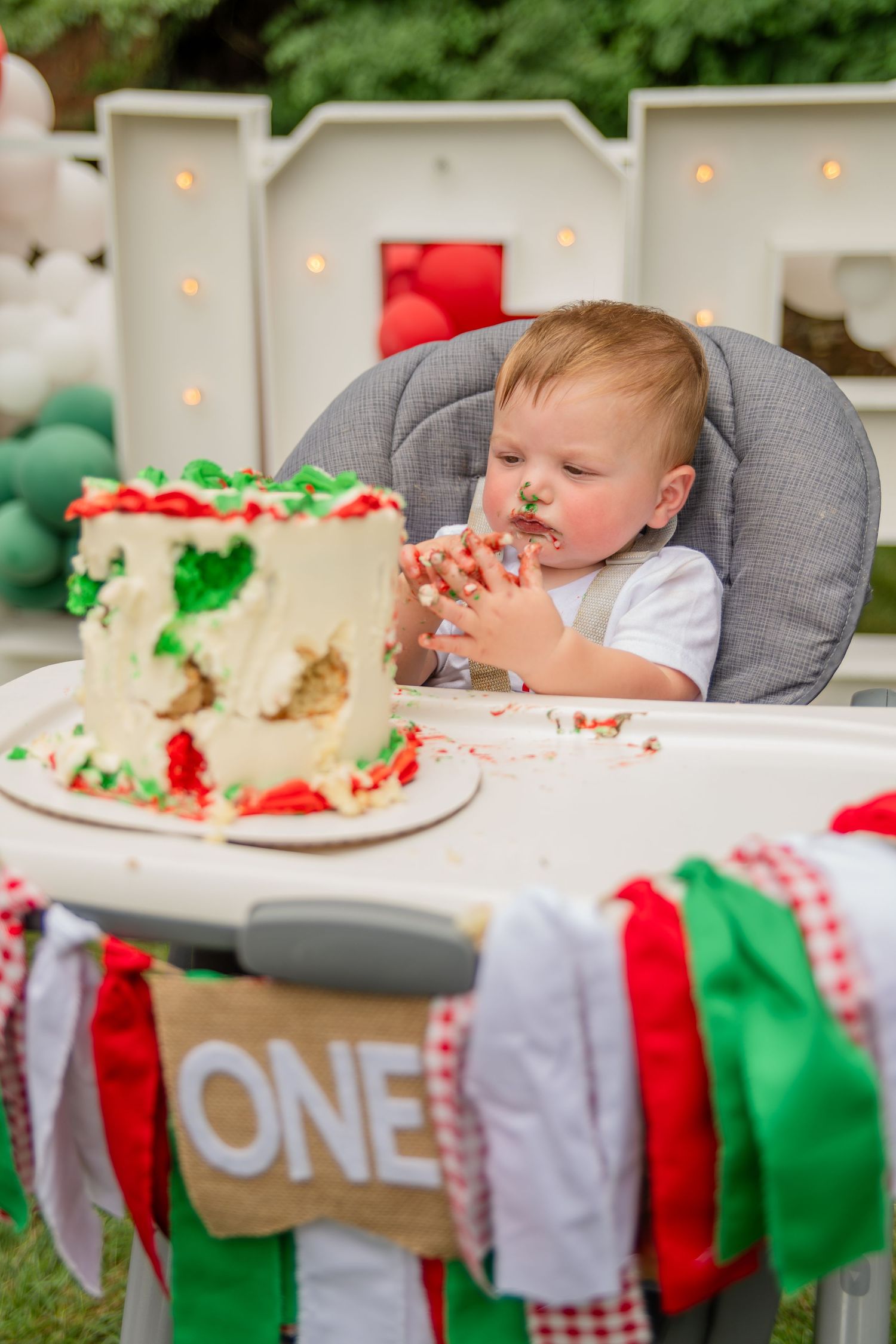 Baby in highchair eagerly eating birthday cake with red and green party decorations.