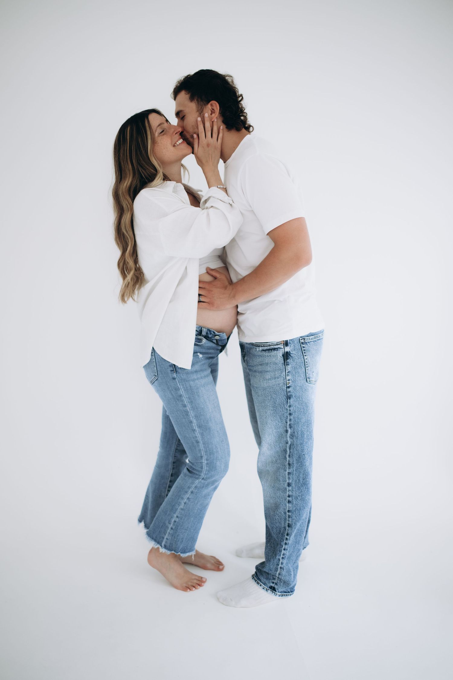 A couple in white tops and blue jeans share an intimate moment against a white studio background.