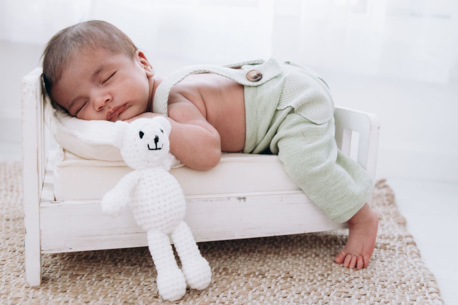Baby sleeping peacefully on small white bed with plush white teddy bear toy on beige carpet.