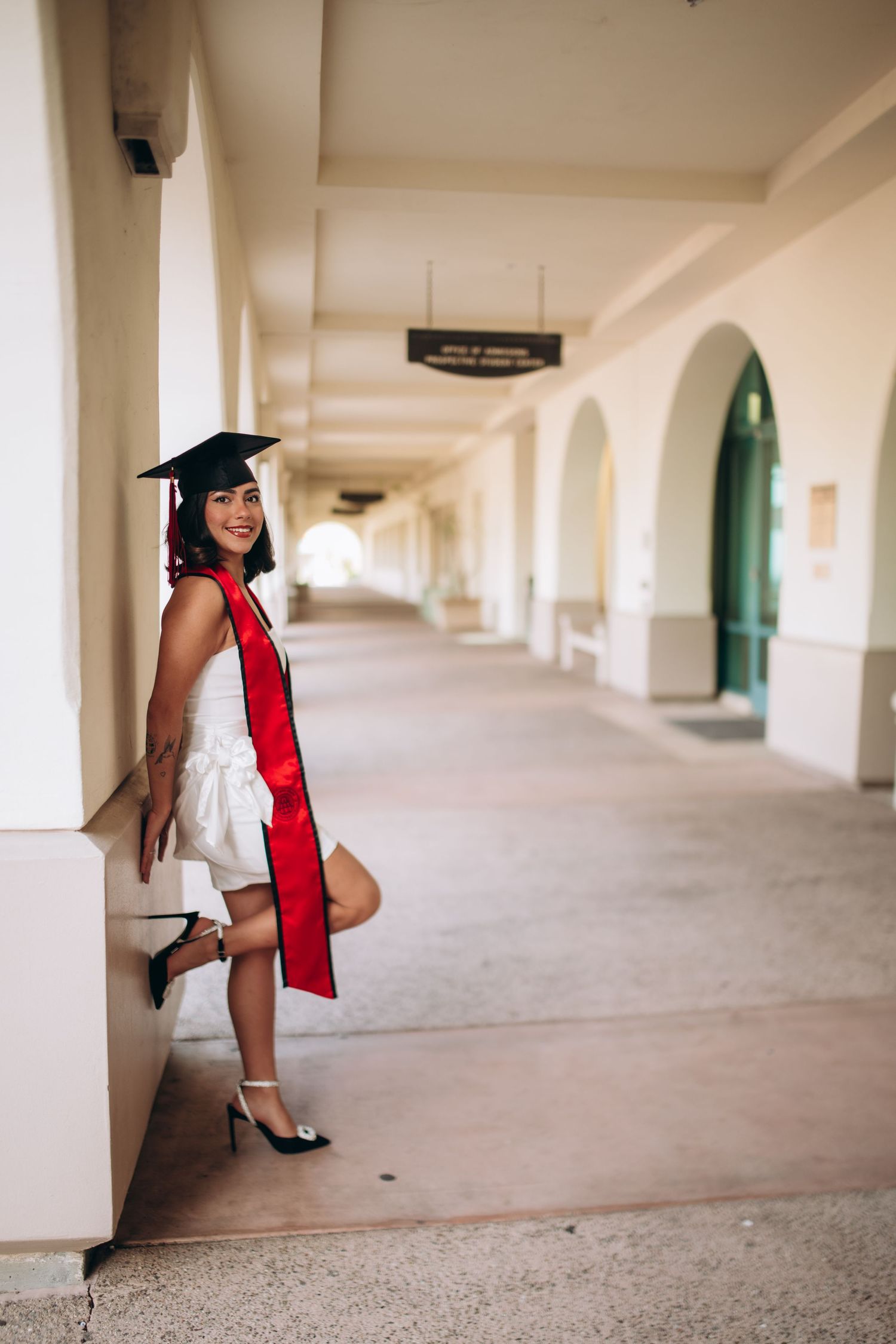 Graduate in white dress and red stole poses in Spanish colonial archway corridor at university.