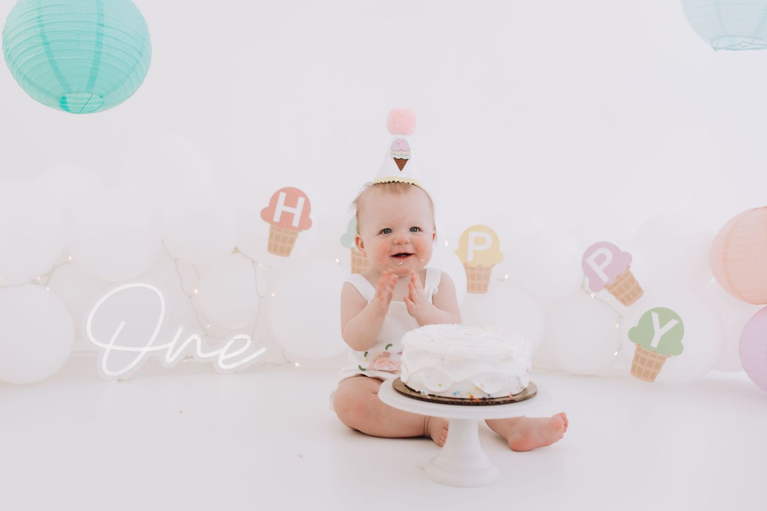 Baby sitting with a white cake during first birthday photoshoot with pastel decorations.