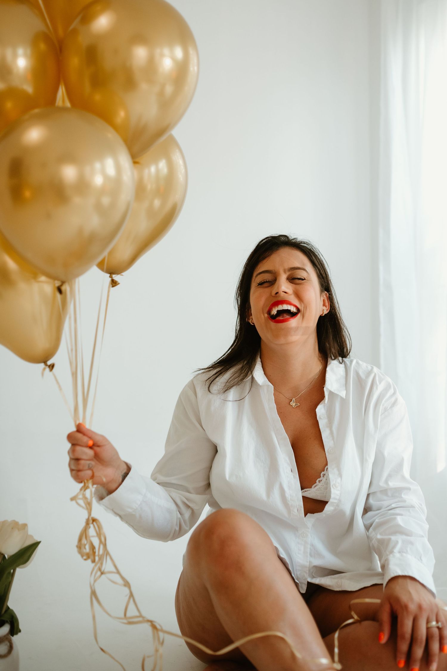 Woman in white button-down shirt laughs while holding gold balloons.