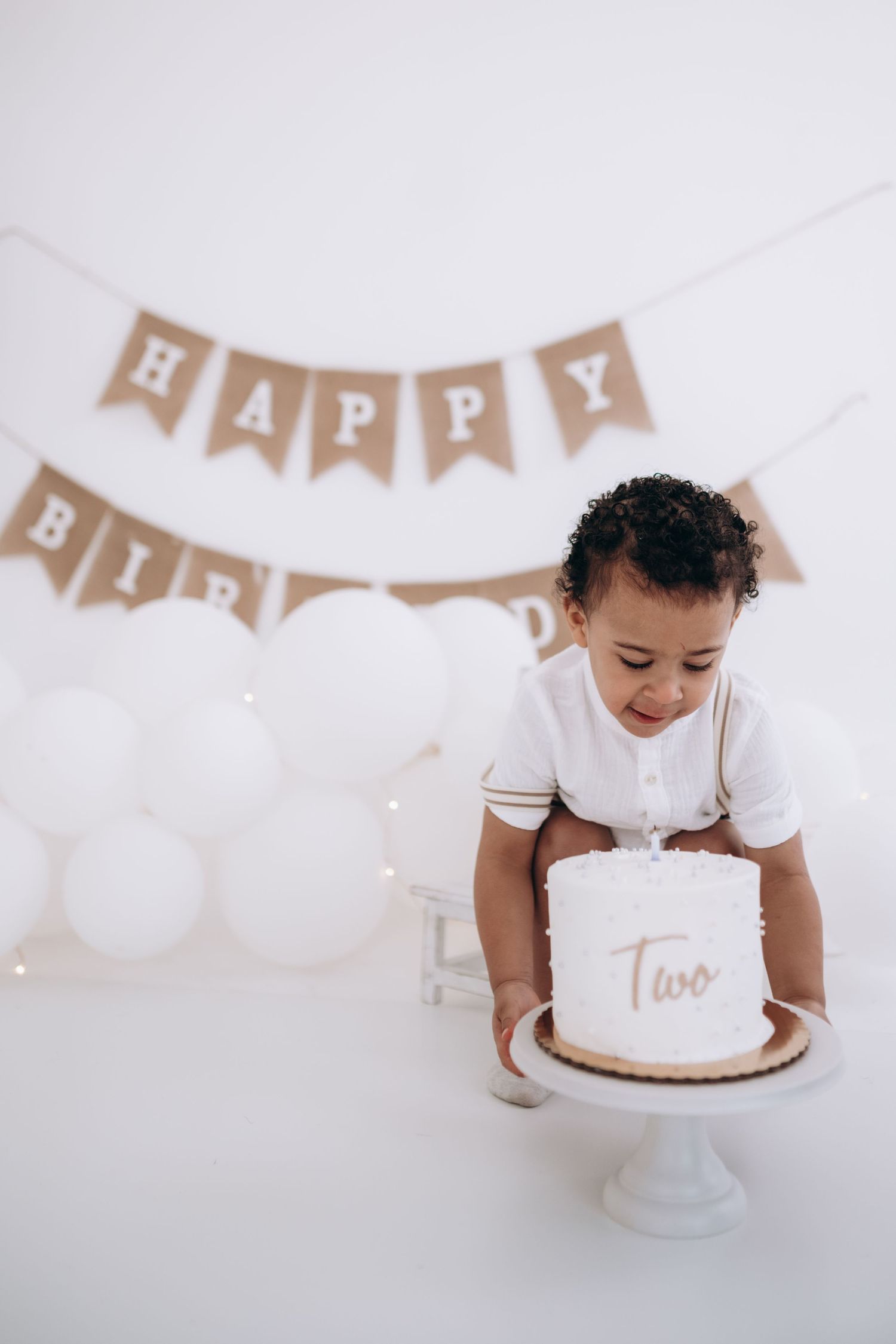 cake smash photography, Toddler in white outfit with birthday cake and Happy Birthday banner decoration.