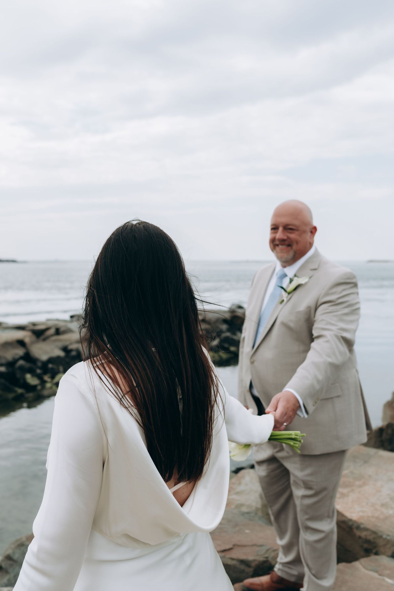 Romantic beach photo session with a couple in white and beige formal attire posing on rocky coastline.