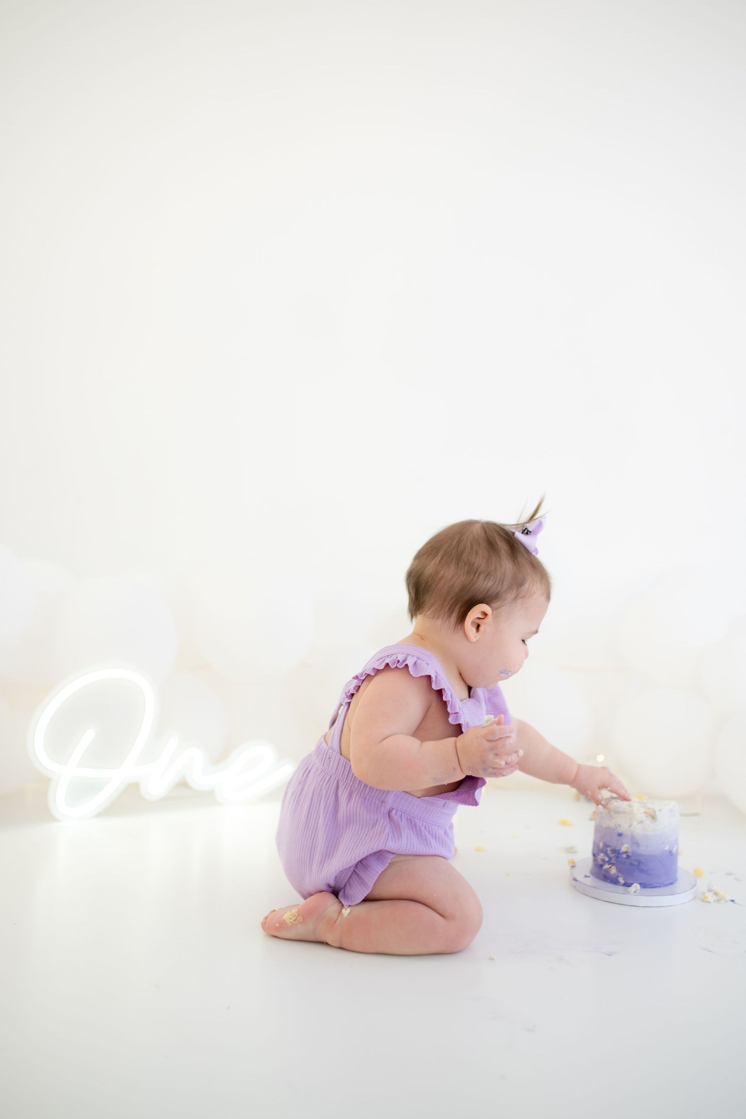cake smash photography,  Baby in purple romper reaching for cake during first birthday photoshoot.