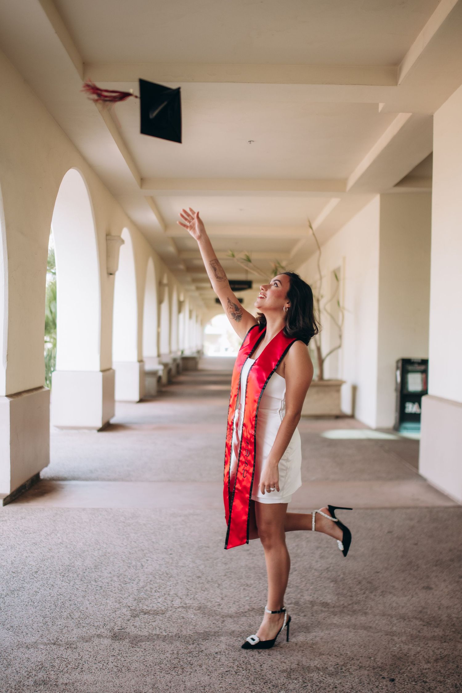 Graduate tosses cap in white dress and red stole in university hallway.