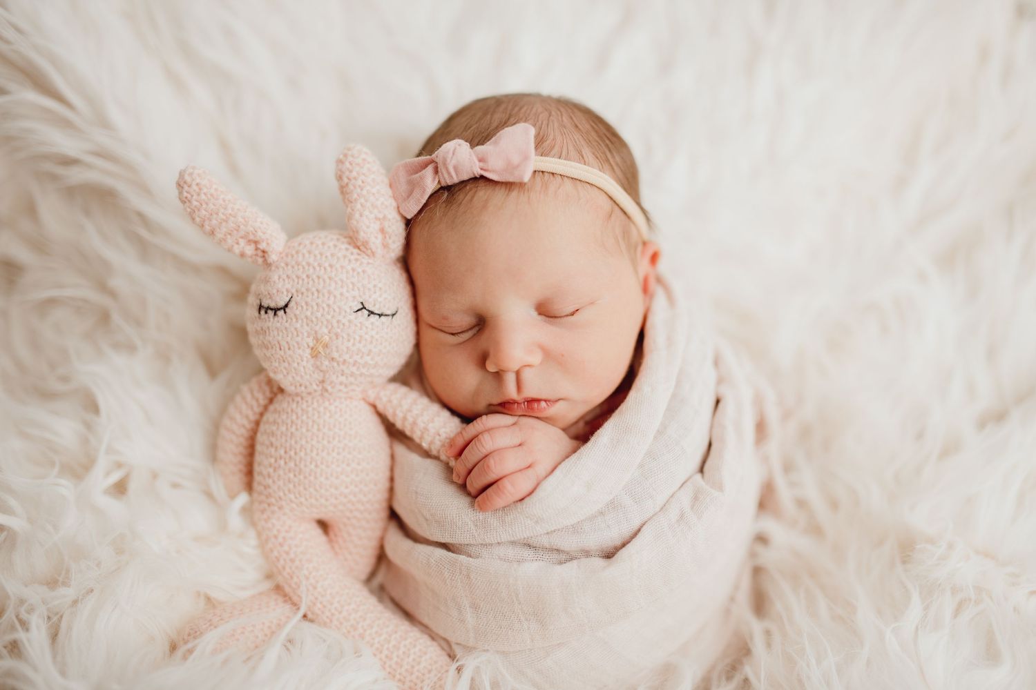 Sleeping newborn wrapped in white swaddle rests beside plush bunny toy on fluffy white blanket.