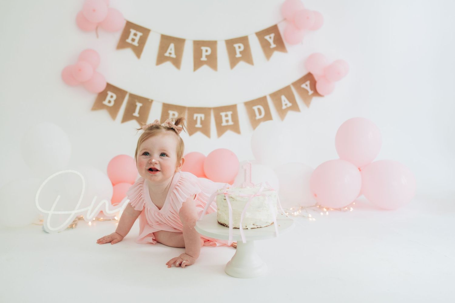 Pink and gold first birthday party setup featuring cake, balloons, and banner decor in soft pastel colors.
