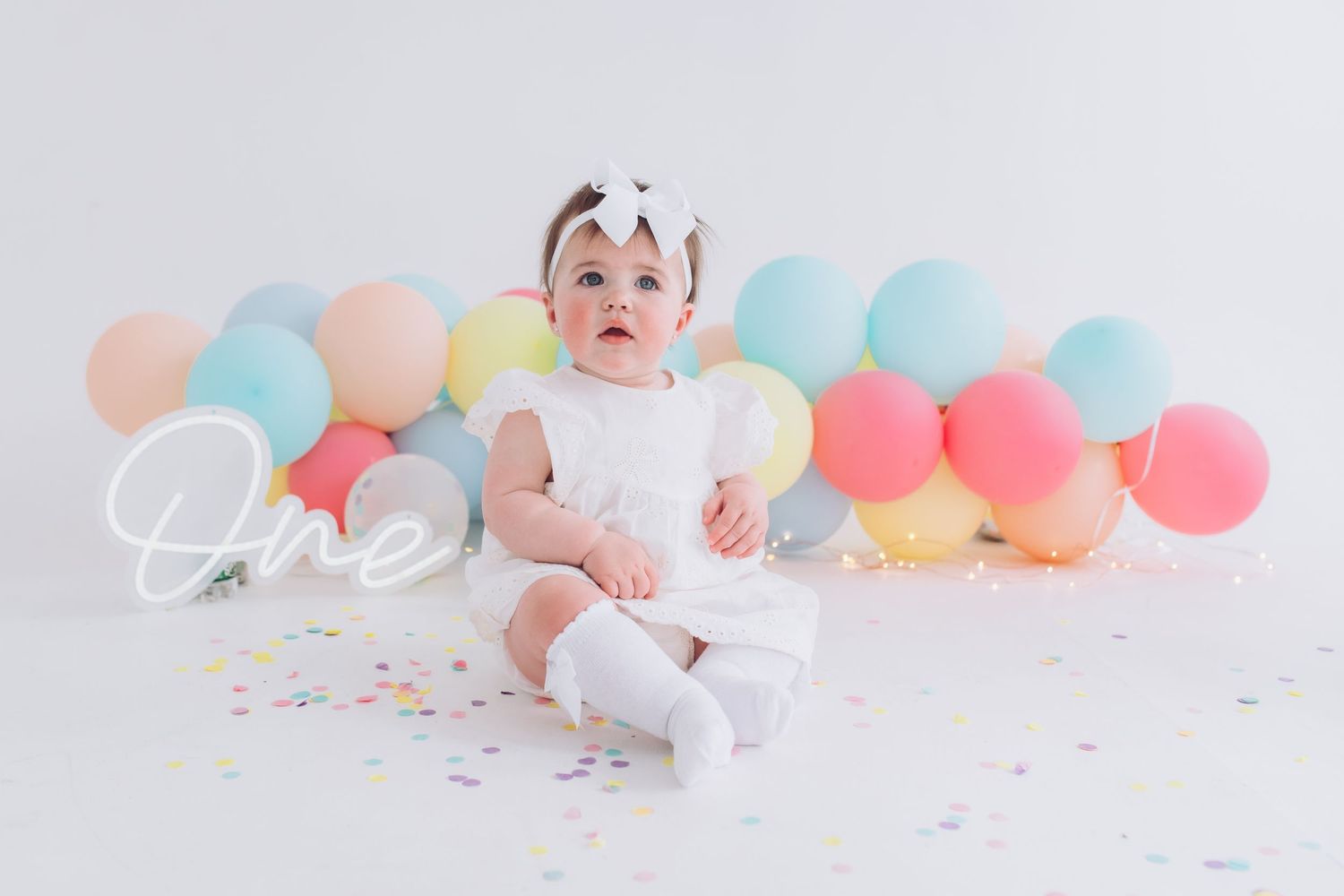 Baby in white outfit sitting on floor with colorful pastel balloons and confetti for milestone photo session.