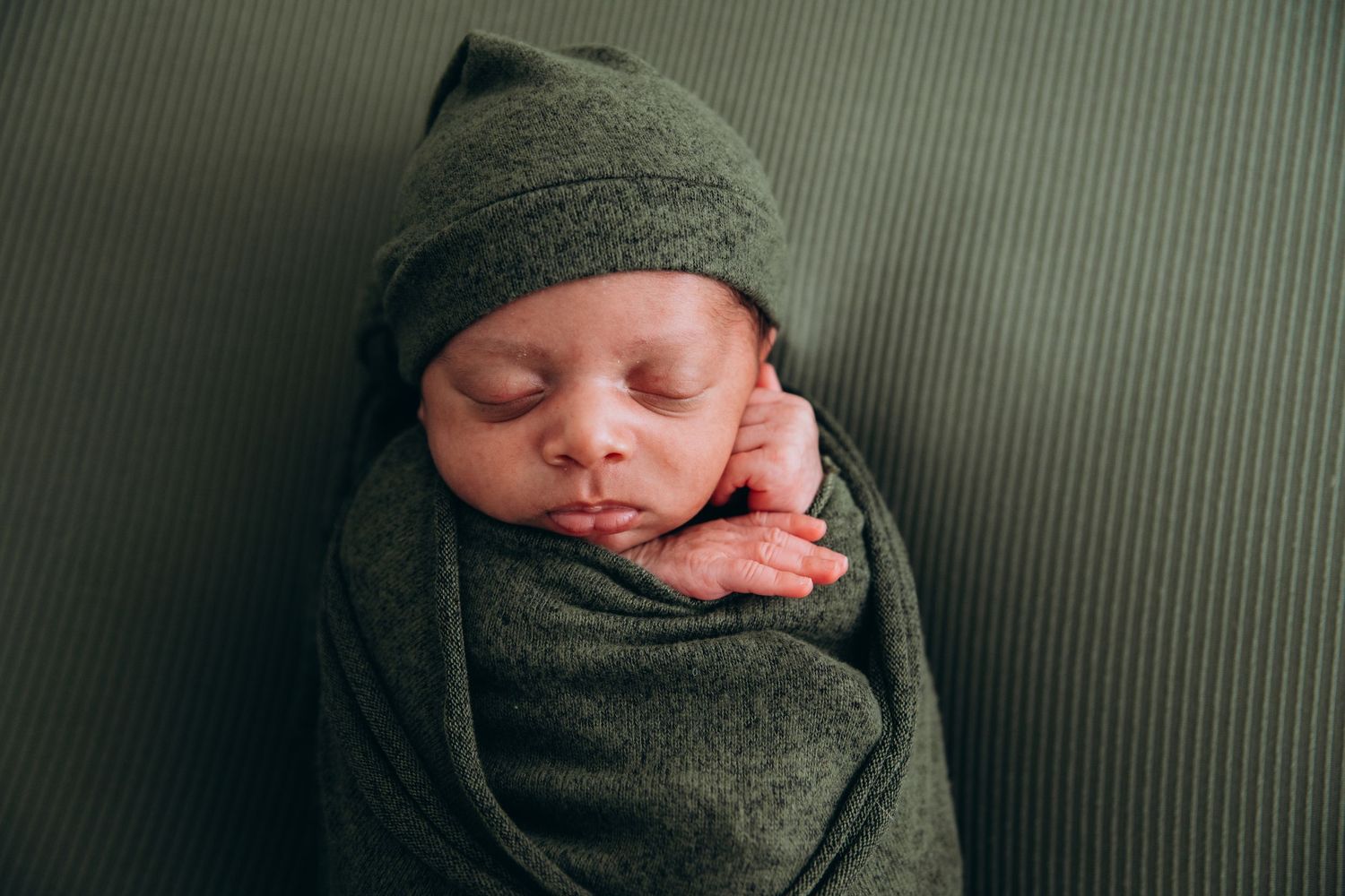 A row of five images showing a sleeping newborn baby swaddled in a dark green blanket against a ribbed green background.