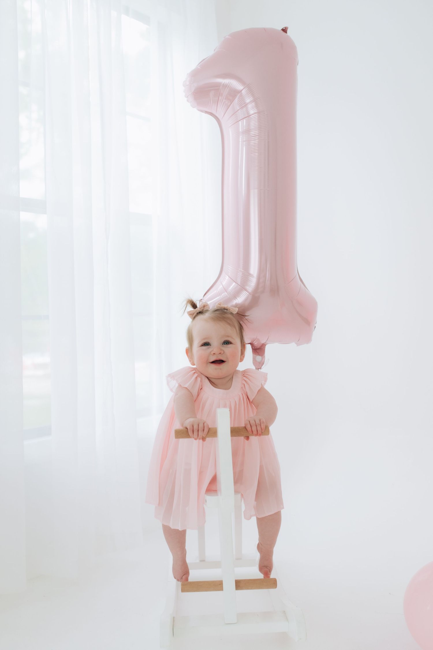 Pink first birthday celebration scene with large number one balloon and small toddler in tulle dress standing on white steps.