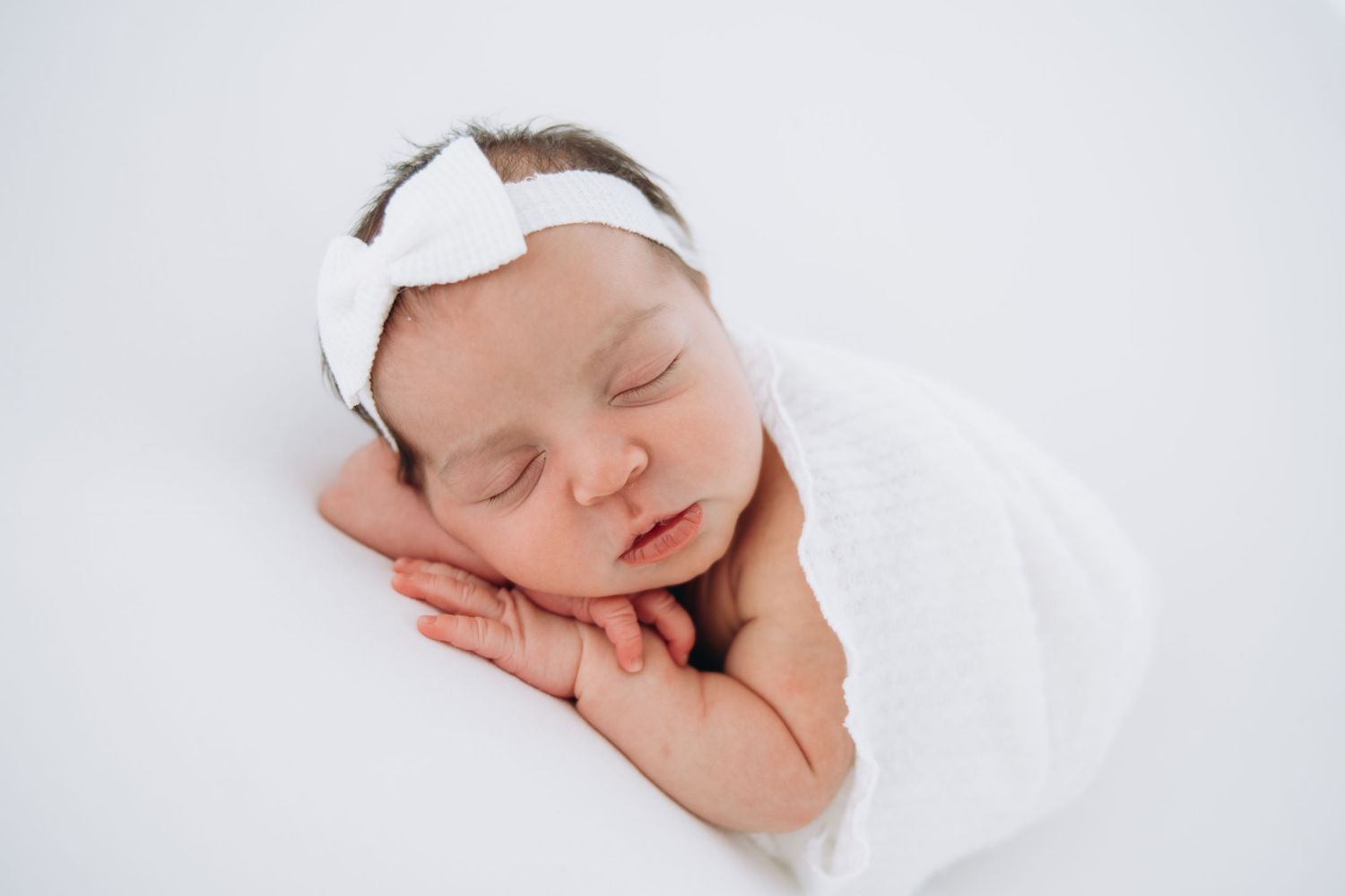 A sleeping newborn swaddled in soft white fabric with a delicate white bow headband rests peacefully.