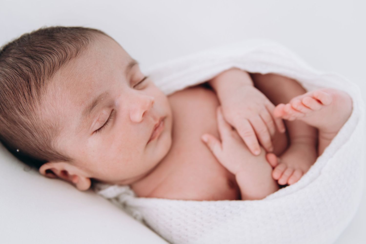 Peaceful sleeping baby wrapped in soft white blanket rests quietly with arms curled against chest.